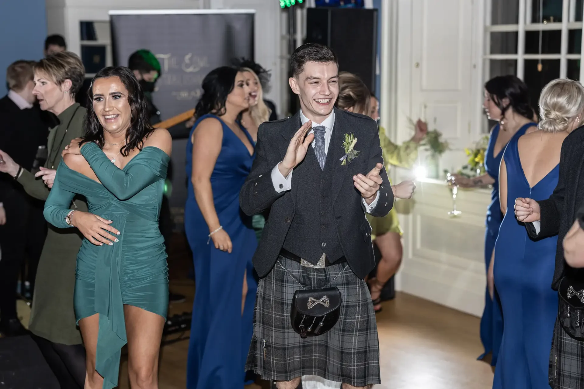 A young man in a kilt smiling as he dances at a lively event, surrounded by elegantly dressed guests.