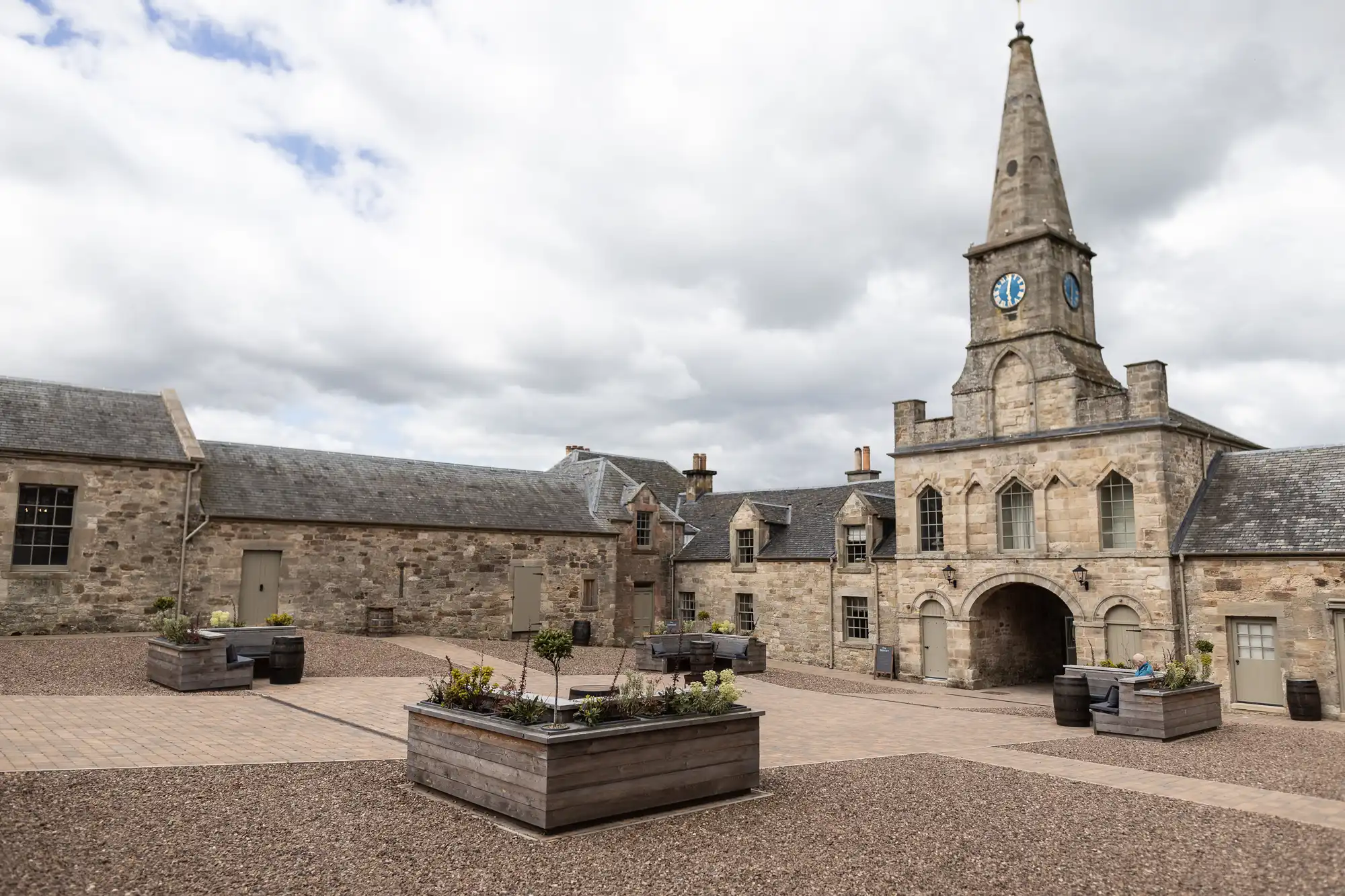 Rosebery Steading courtyard with a central clock tower, surrounded by stone buildings and several large planters with greenery under a cloudy sky.