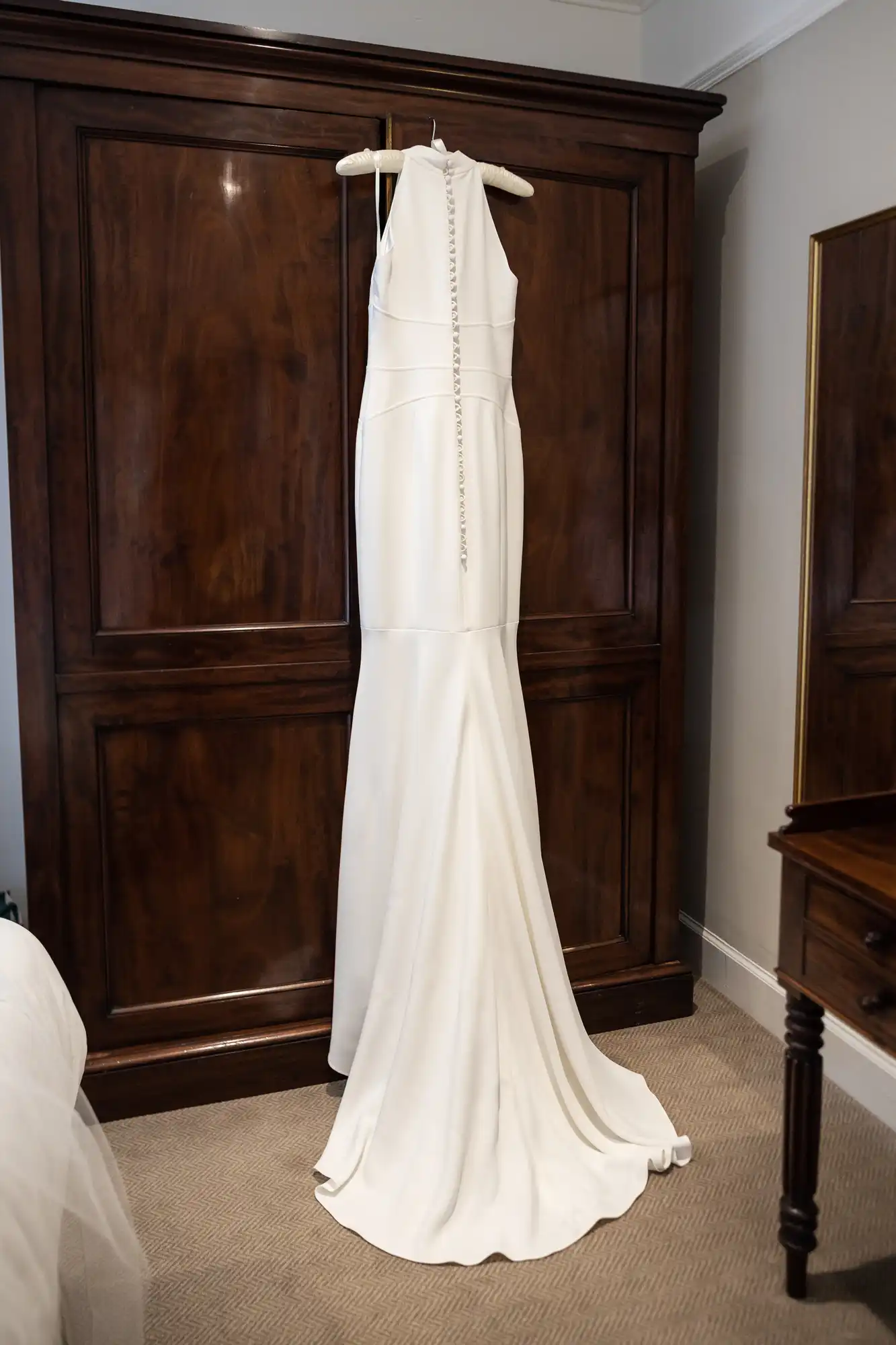 A long, elegant white wedding dress with detailed stitching hangs on a hanger against a dark wooden wardrobe in a room.