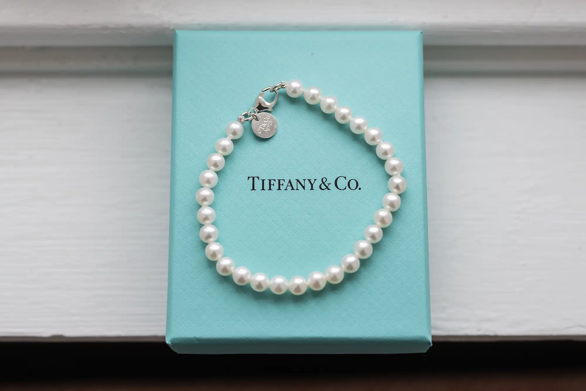 A pearl bracelet with a silver charm rests on a Tiffany & Co. box placed on a white windowsill.