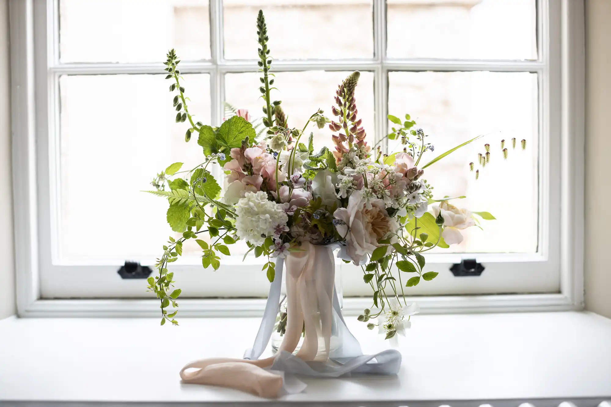 A floral arrangement featuring a mix of flowers and greenery in soft colors, set in front of a bright window on a white sill, adorned with a flowing ribbon.