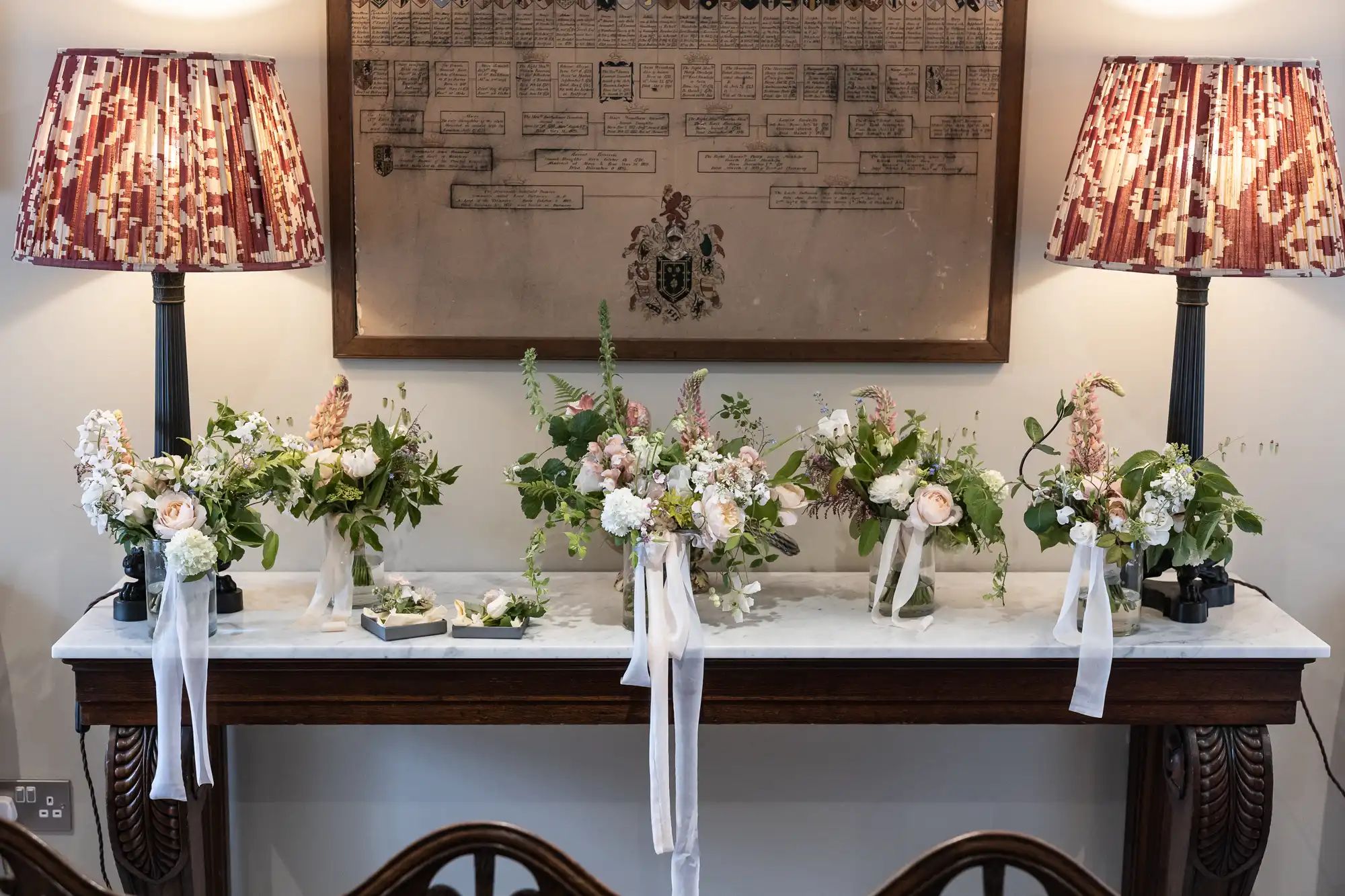 Elegant floral arrangements on a mantelpiece flanked by two lamps, under a framed heraldic emblem.