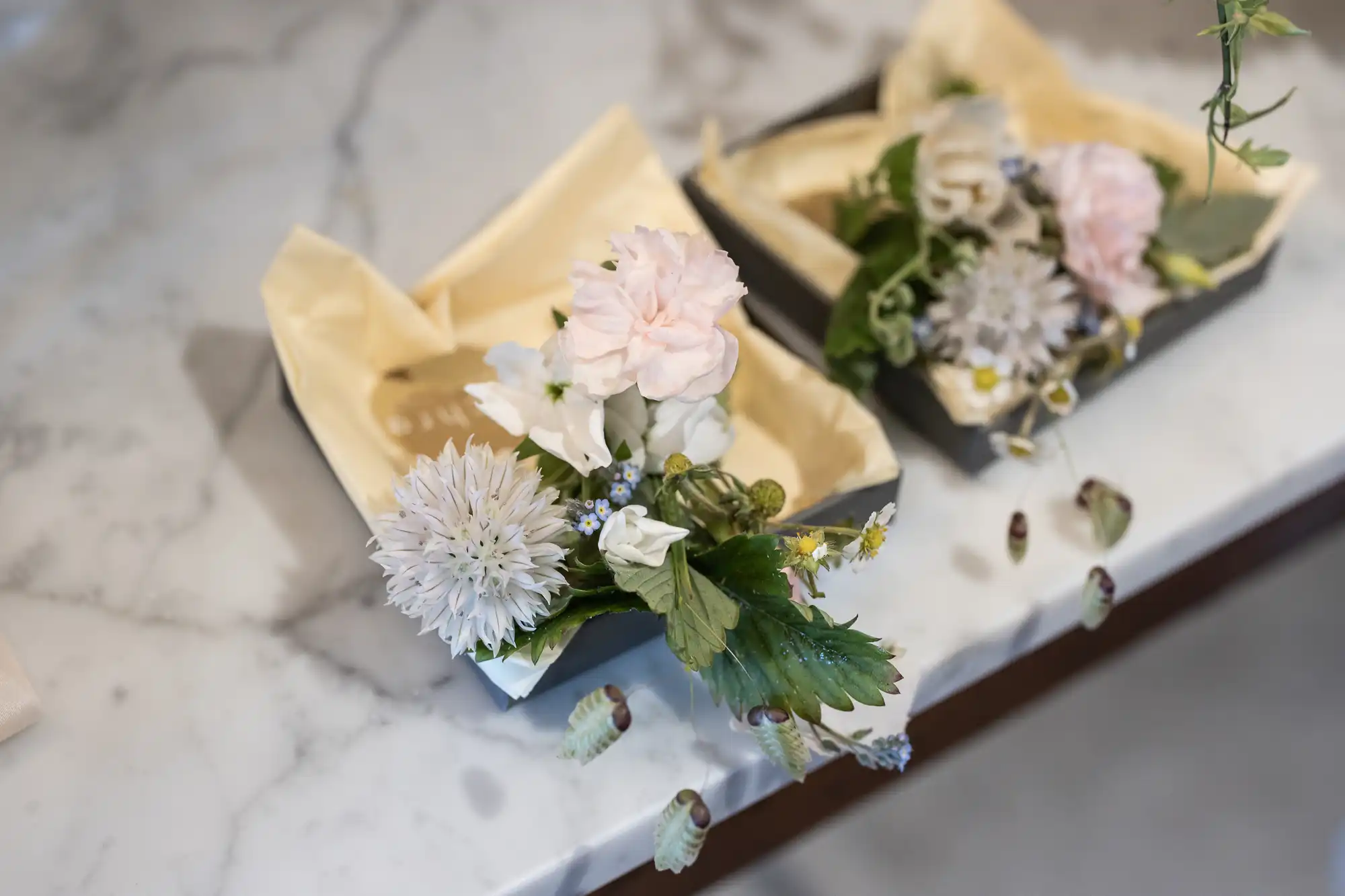 Three floral arrangements in black square containers wrapped with beige paper, displayed on a marble countertop.