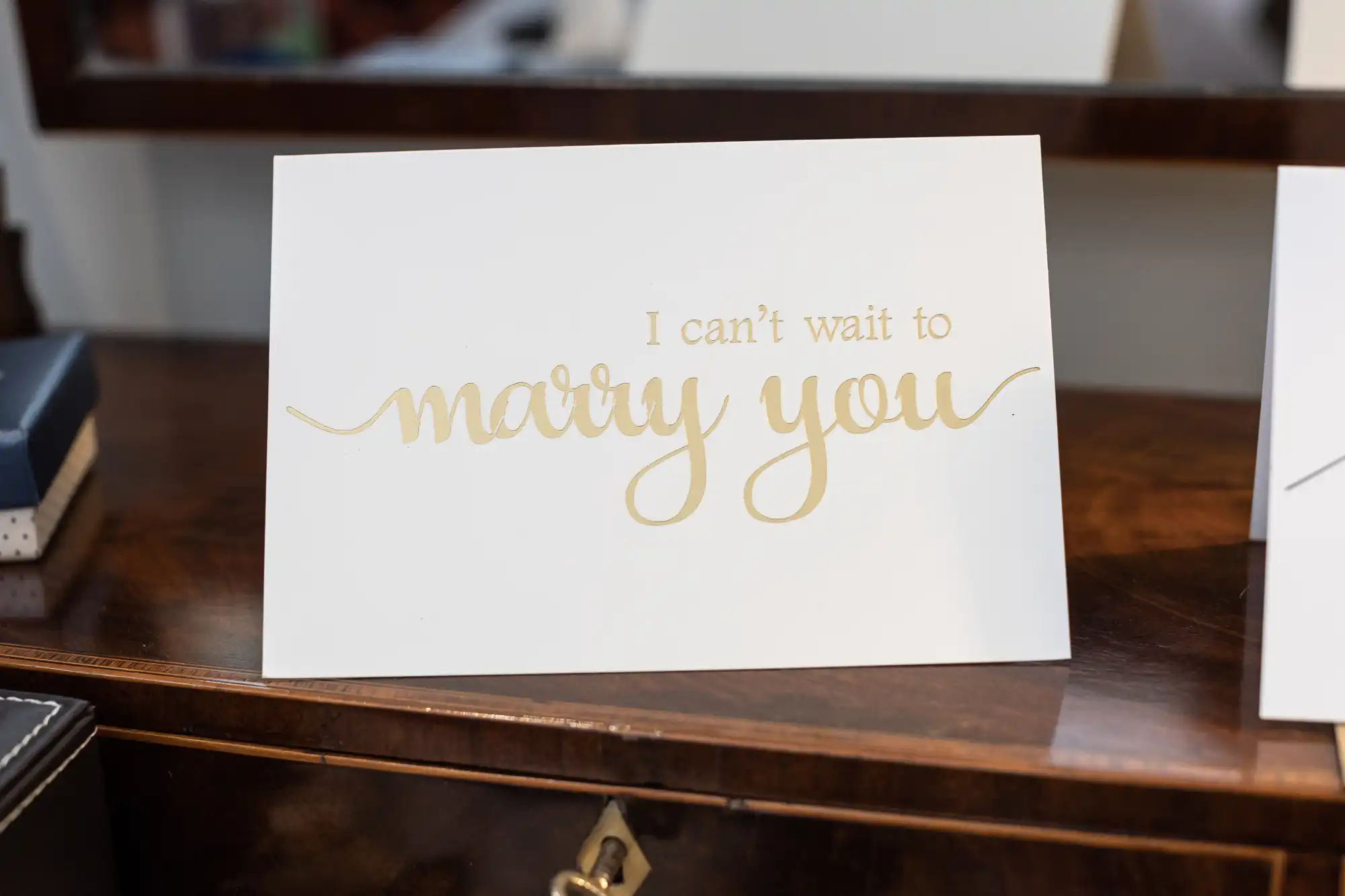 A card with gold cursive writing that says "I can't wait to marry you," placed on a wooden desk.