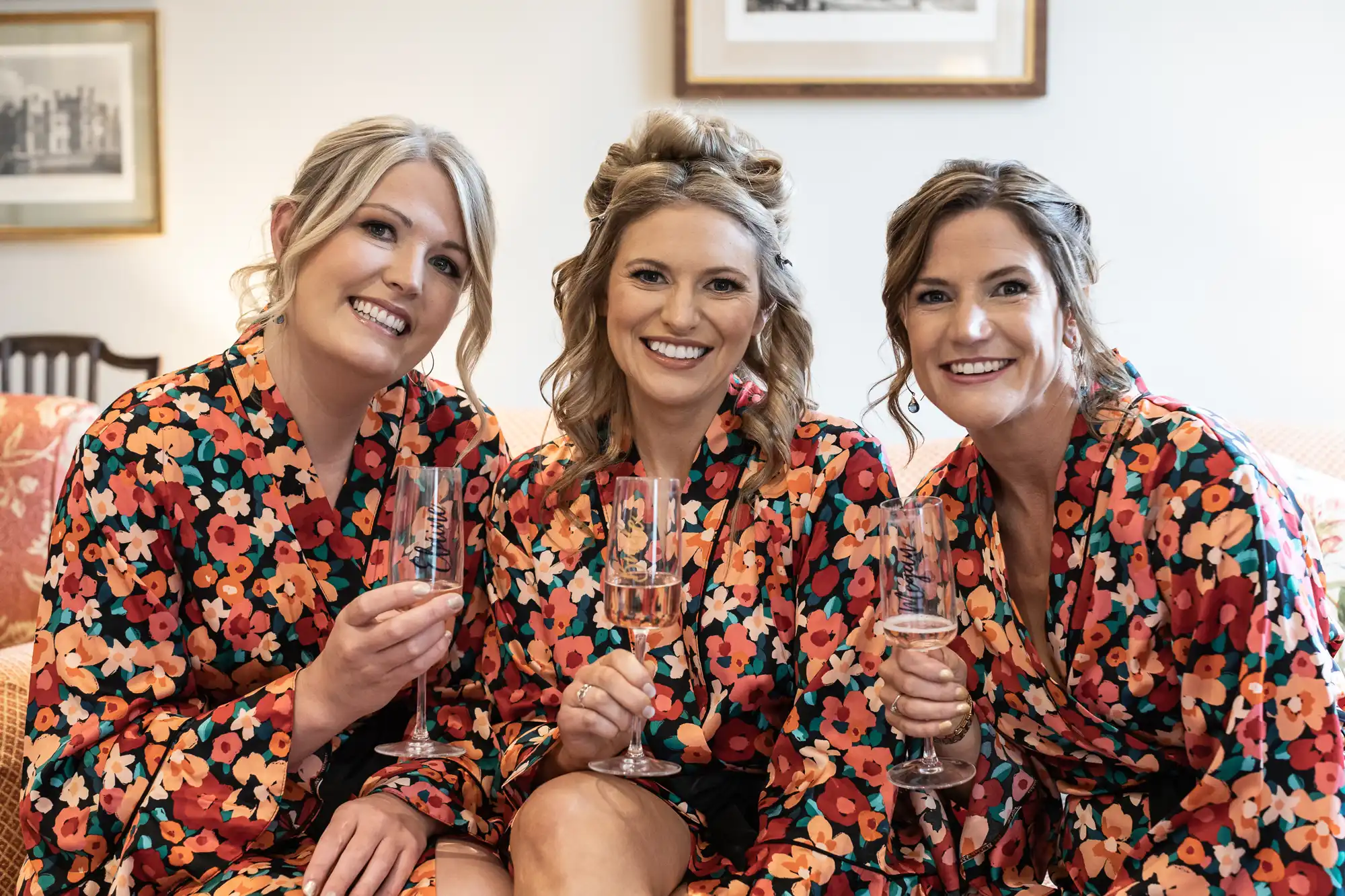 Three women in matching floral robes smiling and holding champagne glasses indoors.