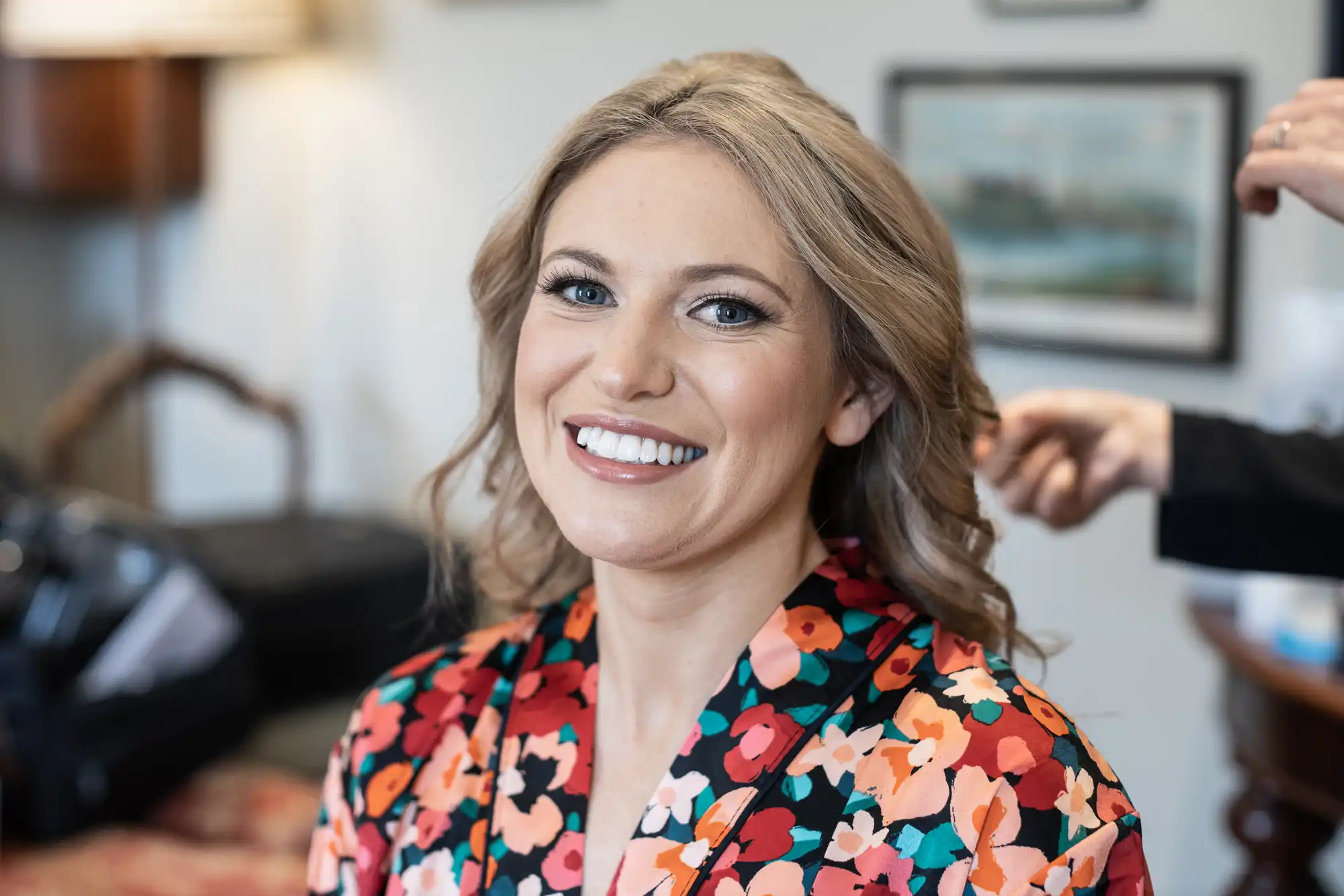 A smiling woman with blonde hair wearing a floral blouse gets her hair styled by a person in the background.