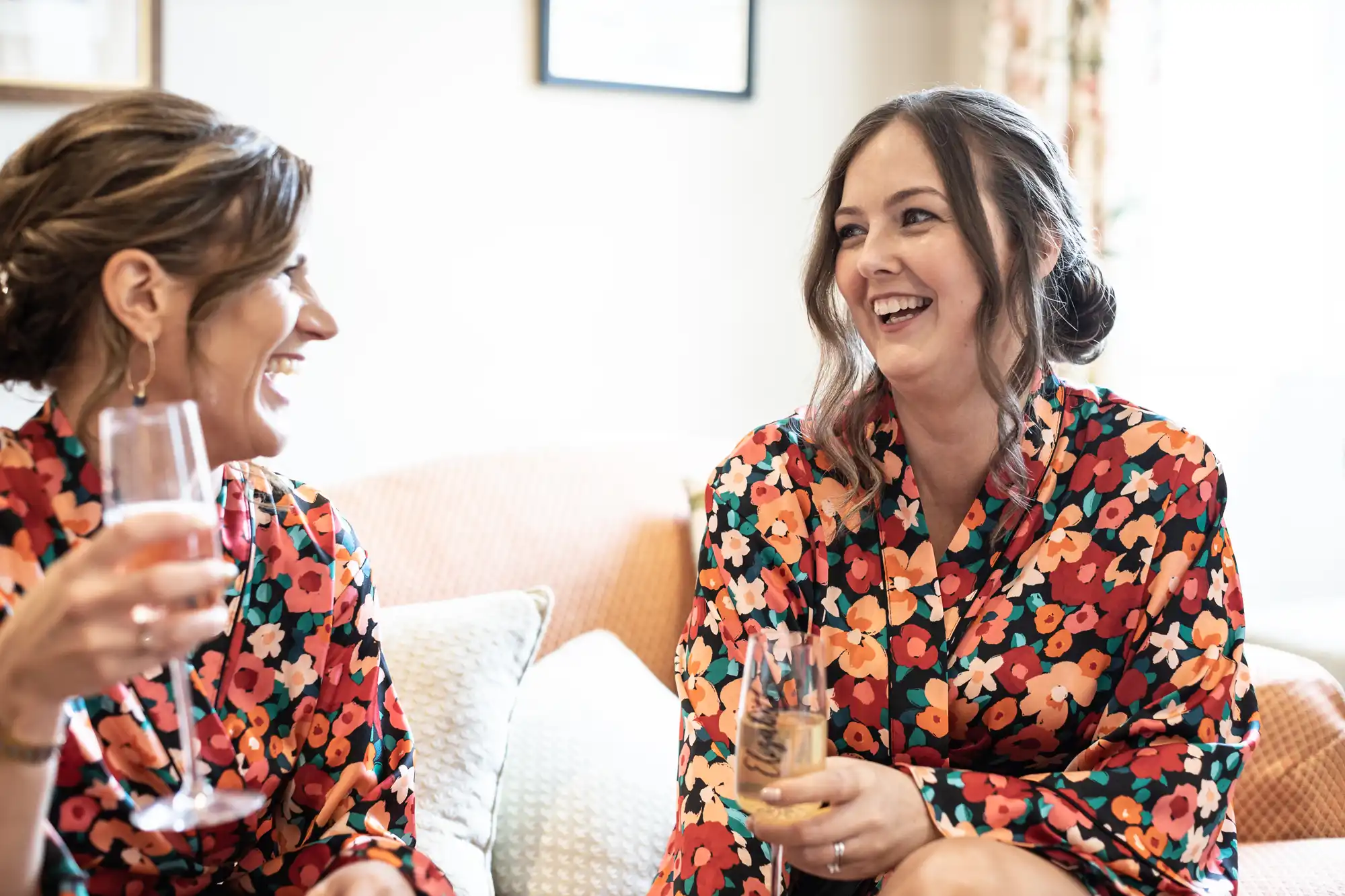 Two women laughing and holding glasses of wine, sitting on a sofa, wearing floral dresses in a well-lit living room.