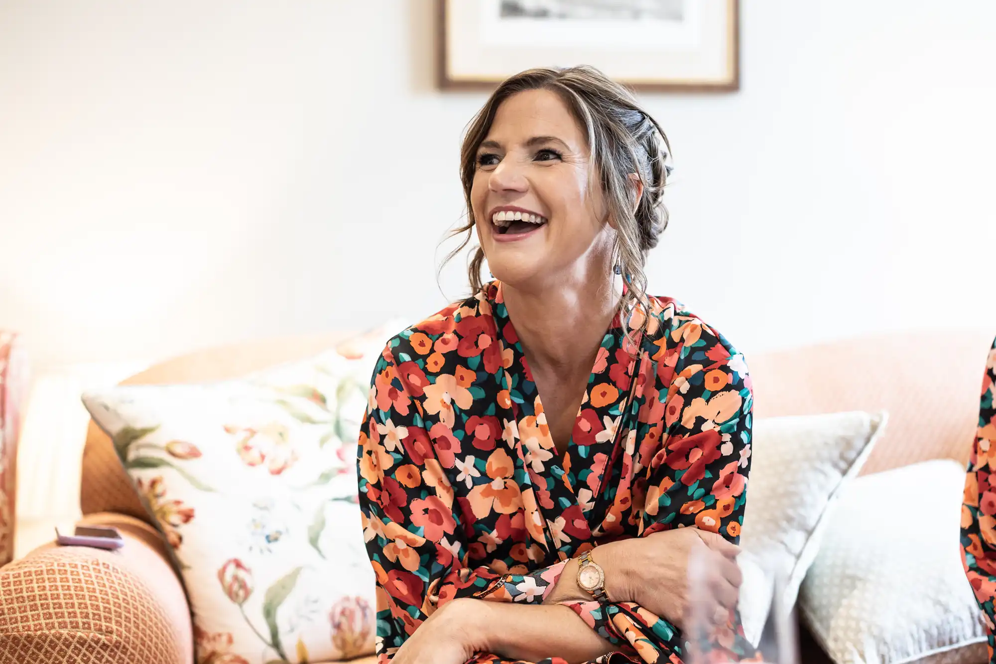 A woman in a floral dress laughs joyfully, sitting on a couch in a warmly lit living room.