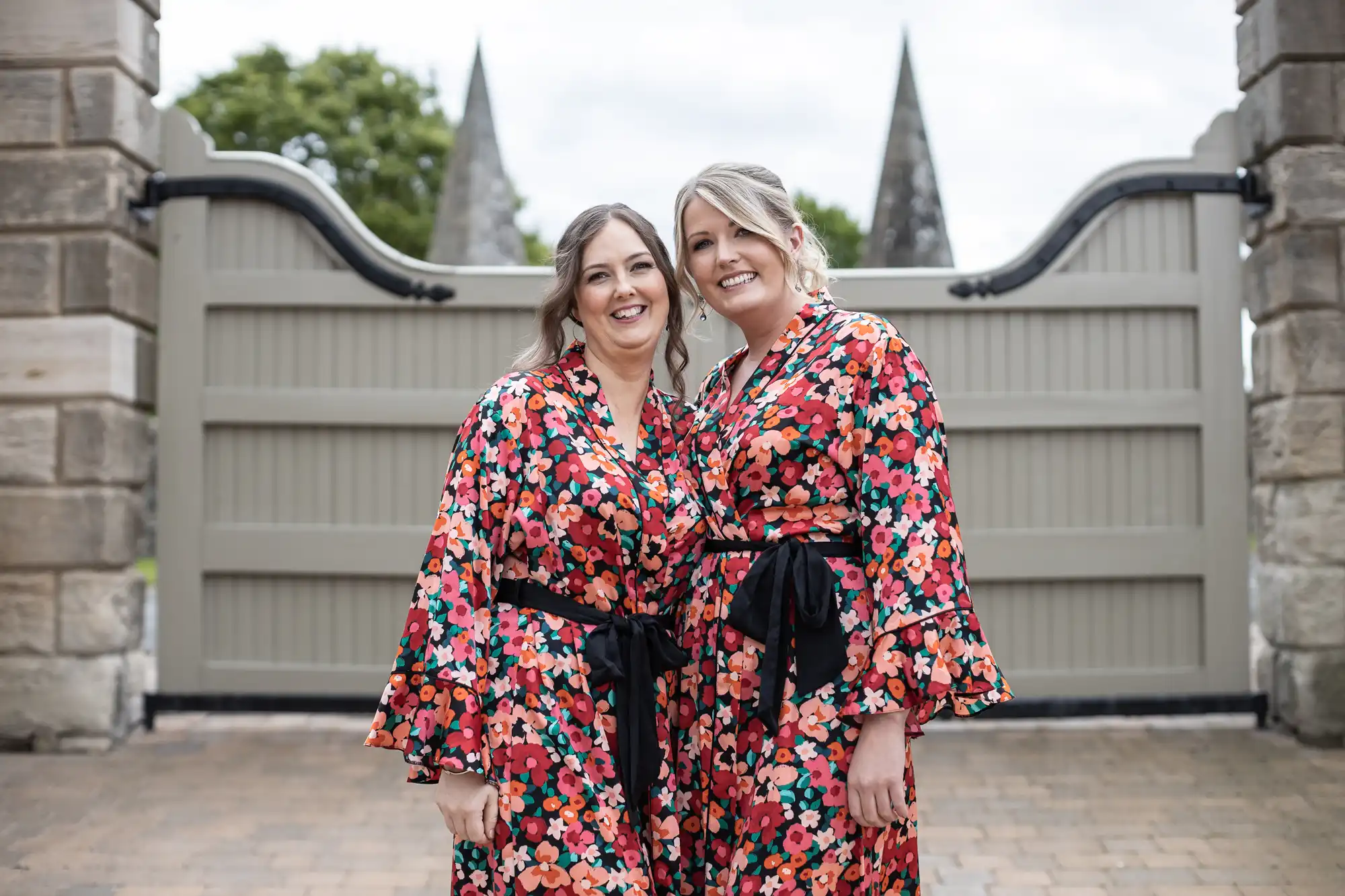 Two women in floral robes smiling and standing arm-in-arm in front of a gate with pointed tops, possibly at a formal event.