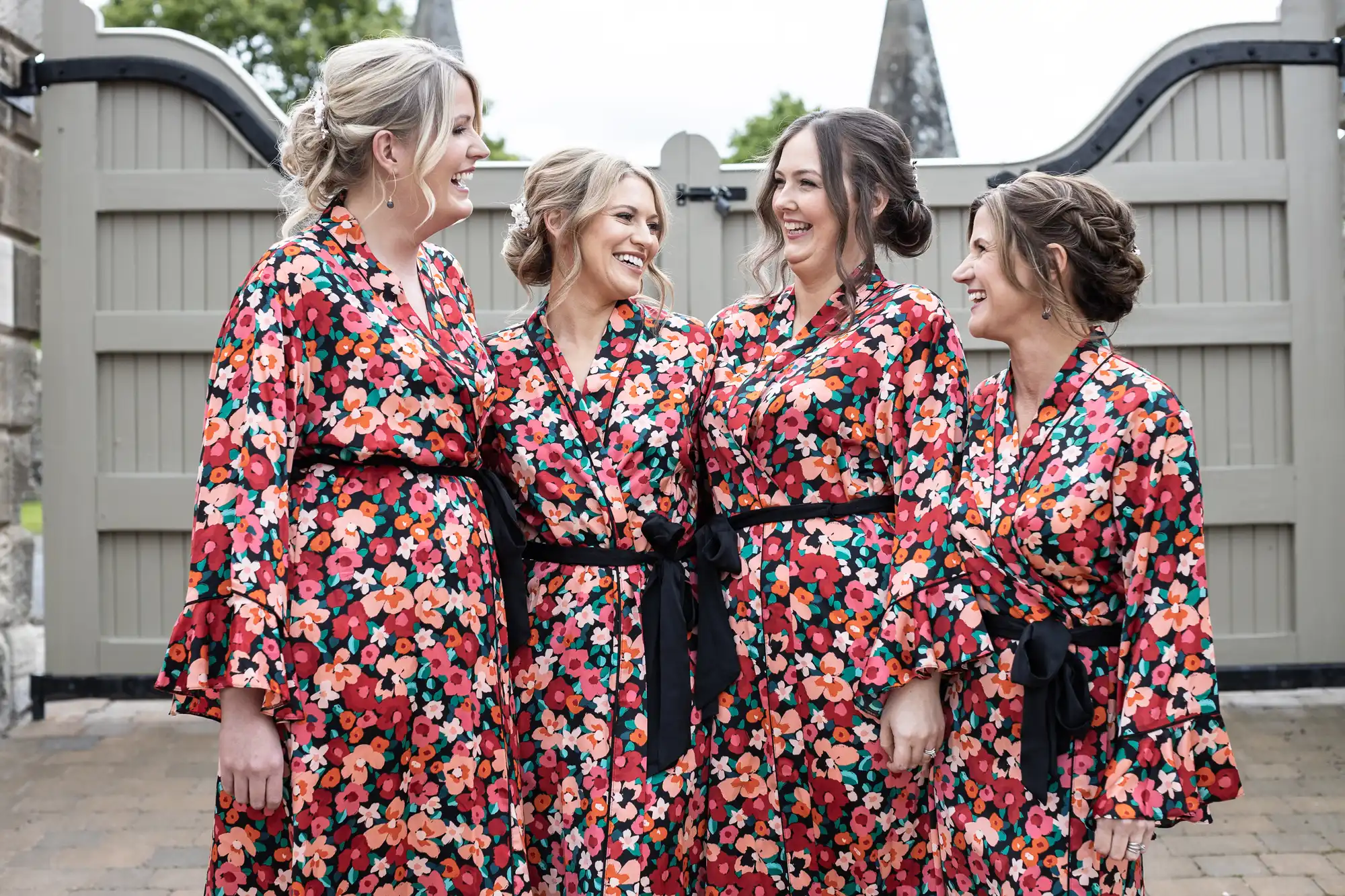 Four women in matching floral robes laugh together in front of a fence with spear-top designs.