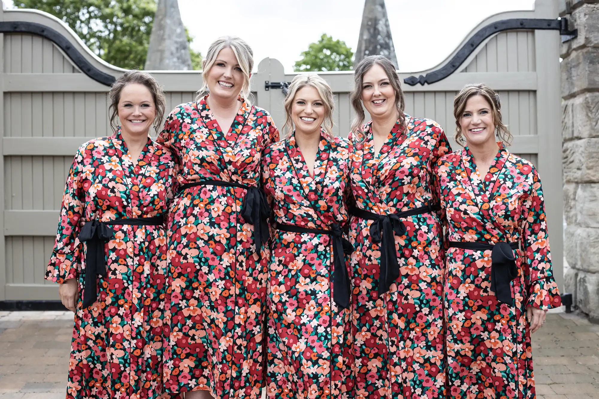 Five women in matching floral dresses smiling in front of a stone fence with castle-like spires.
