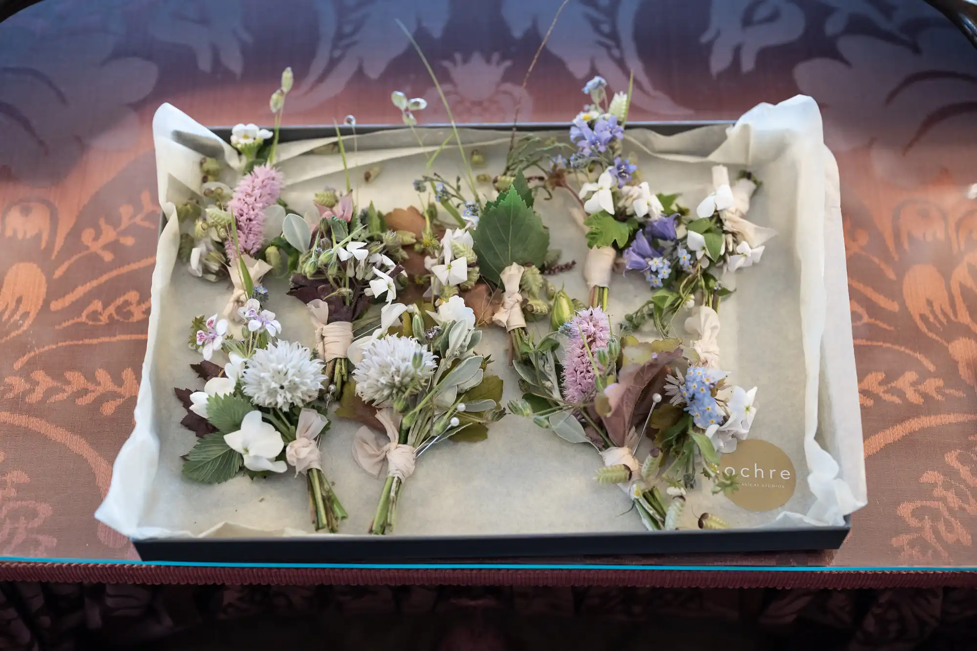 A tray with an assortment of fresh flowers and leaves on parchment paper, presented on a glass surface displaying a reflection.
