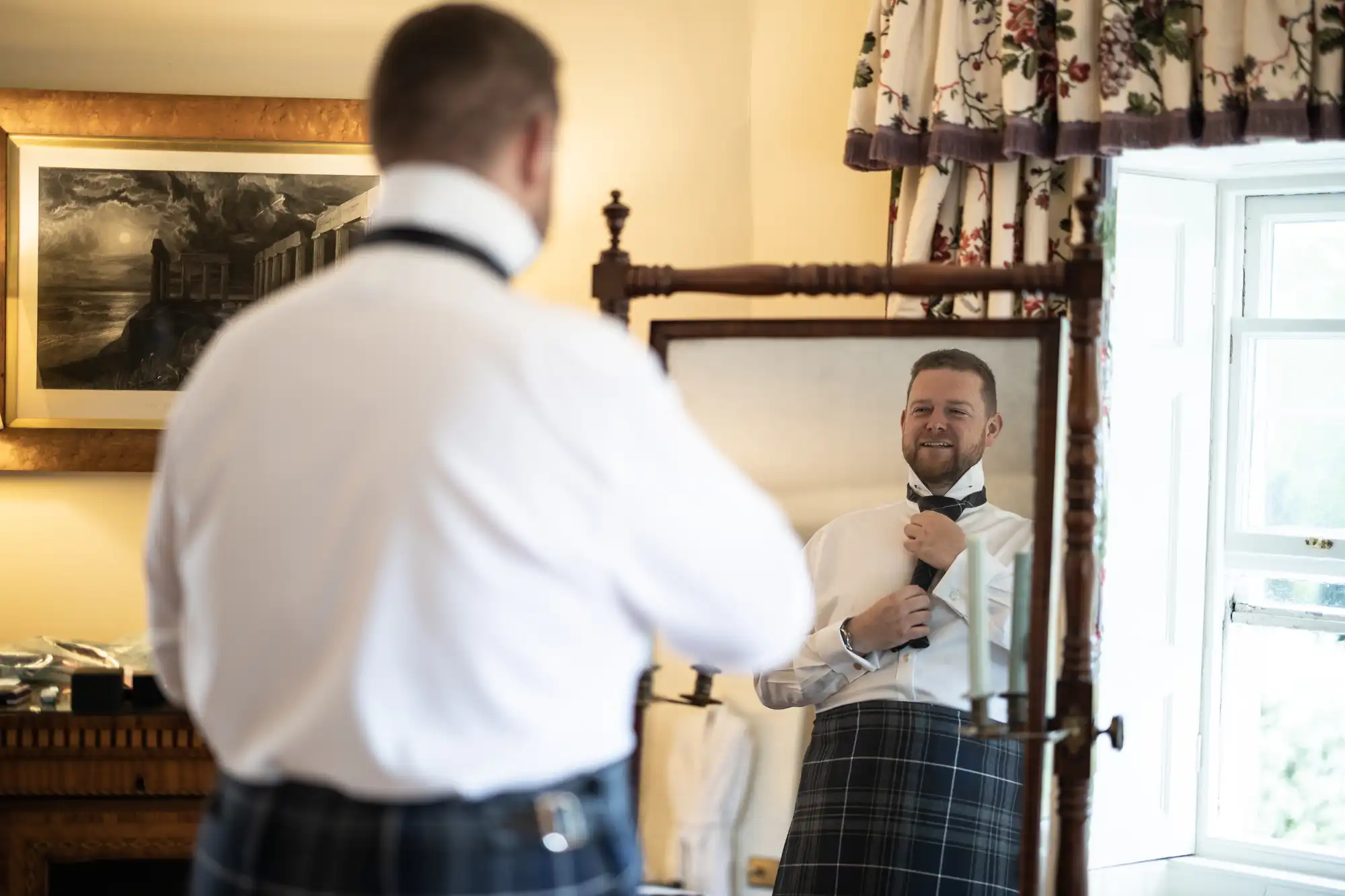 A man in a white shirt and tartan kilt adjusts his tie while smiling at his reflection in a mirror in a warmly lit room.