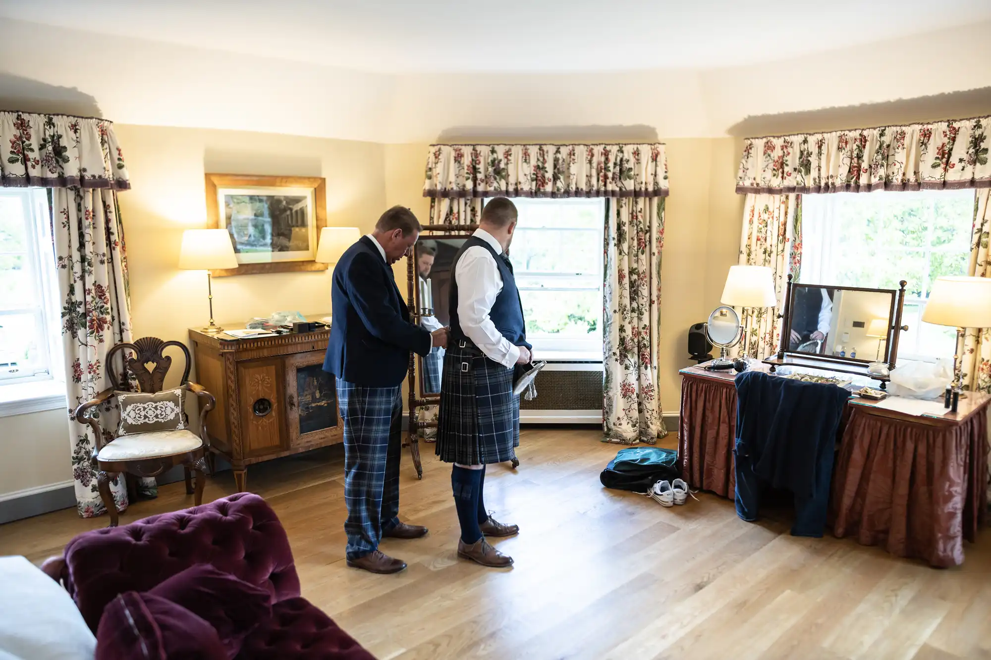 Two men in kilts adjust attire in a traditional room with floral curtains and antique furniture.