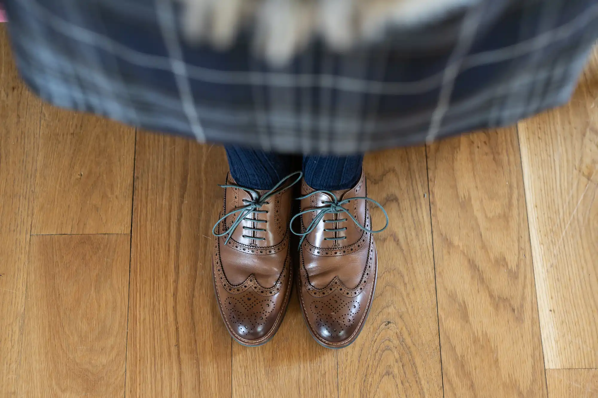 A person standing on a wooden floor wearing brown brogue shoes and blue jeans, with the edge of a plaid shirt visible.