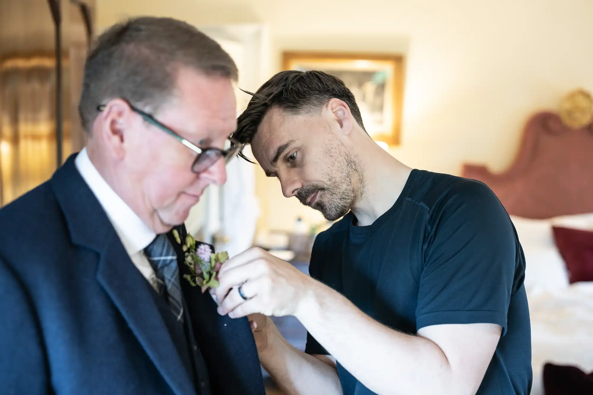 A man adjusts a boutonniere on the lapel of an older man’s suit jacket in a warmly lit room.