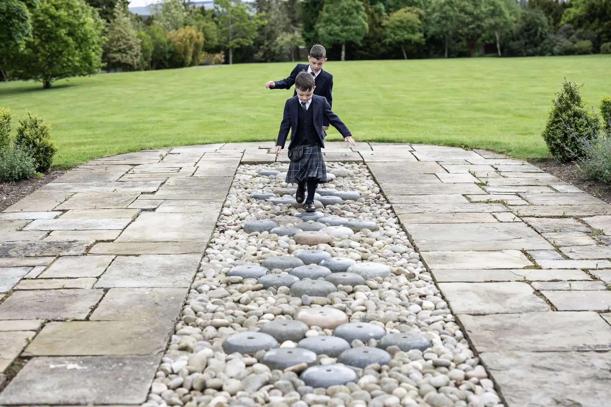 Two children in school uniforms playing on a stone path lined with pebbles and grass in a landscaped garden.