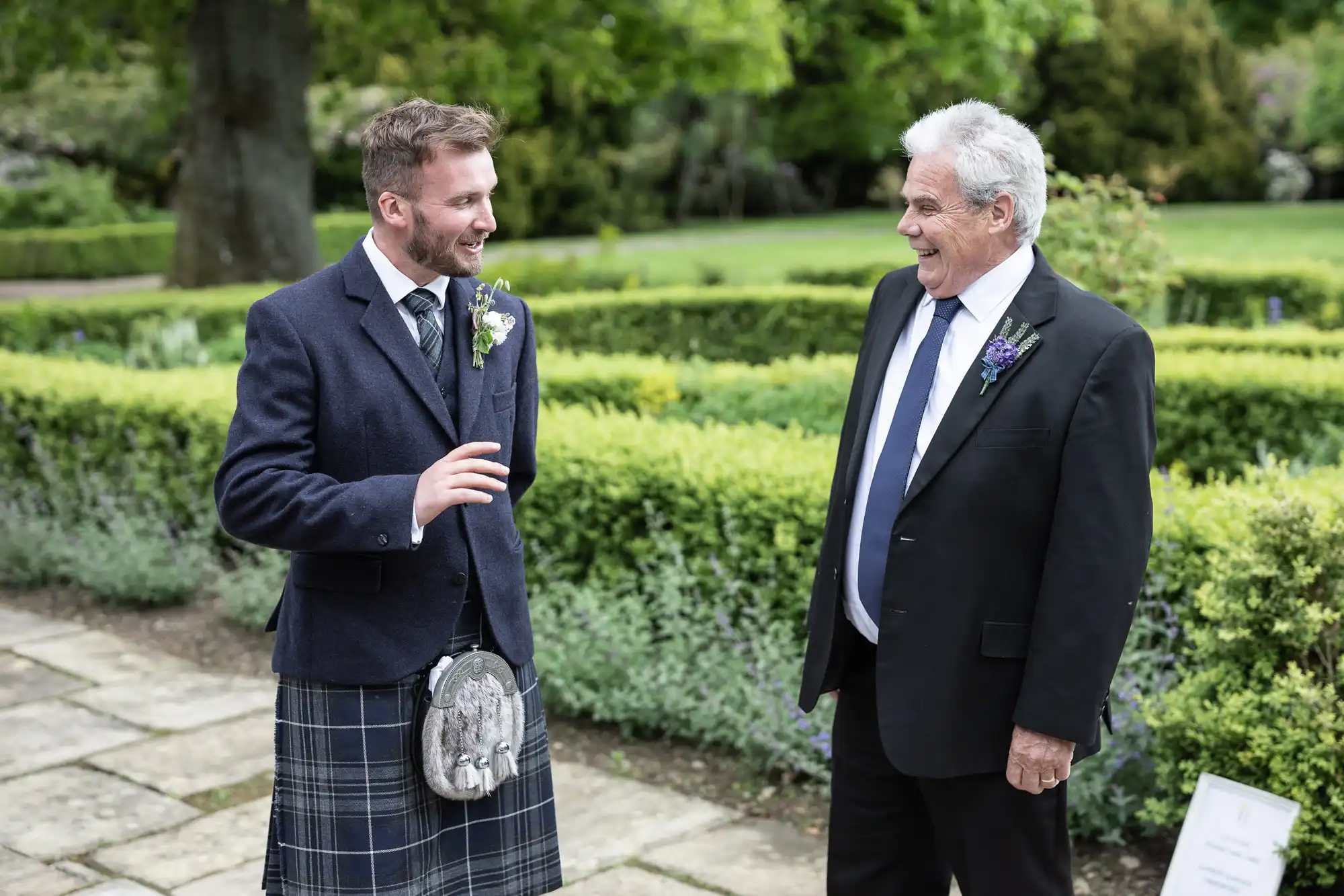 Two men in formal attire, one in a kilt, smiling and talking at an outdoor event with greenery in the background.