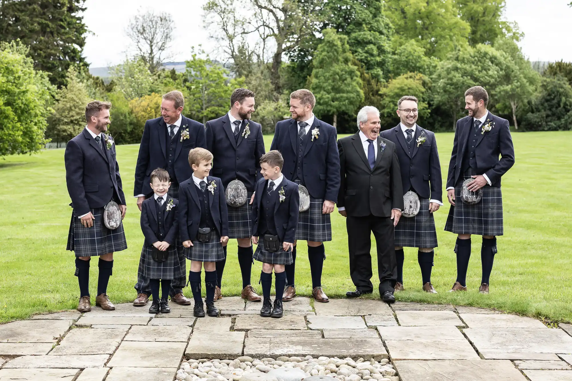 Group of men and boys in traditional Scottish attire with kilts, standing outdoors, smiling on a grassy background.