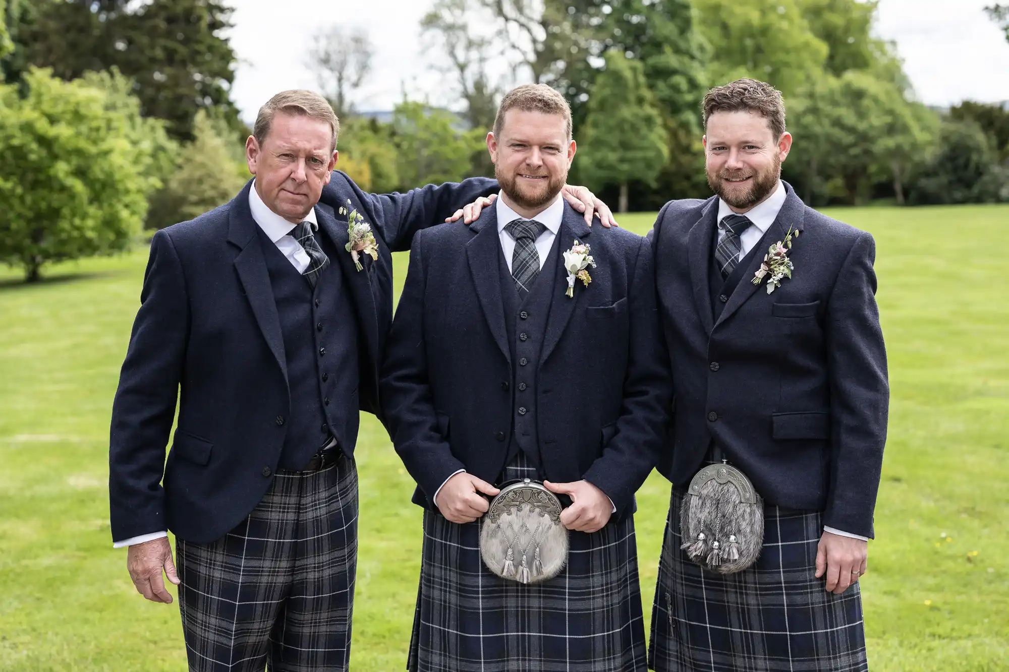 Three men in traditional Scottish kilts and jackets with floral boutonnieres, standing together and smiling in a garden.