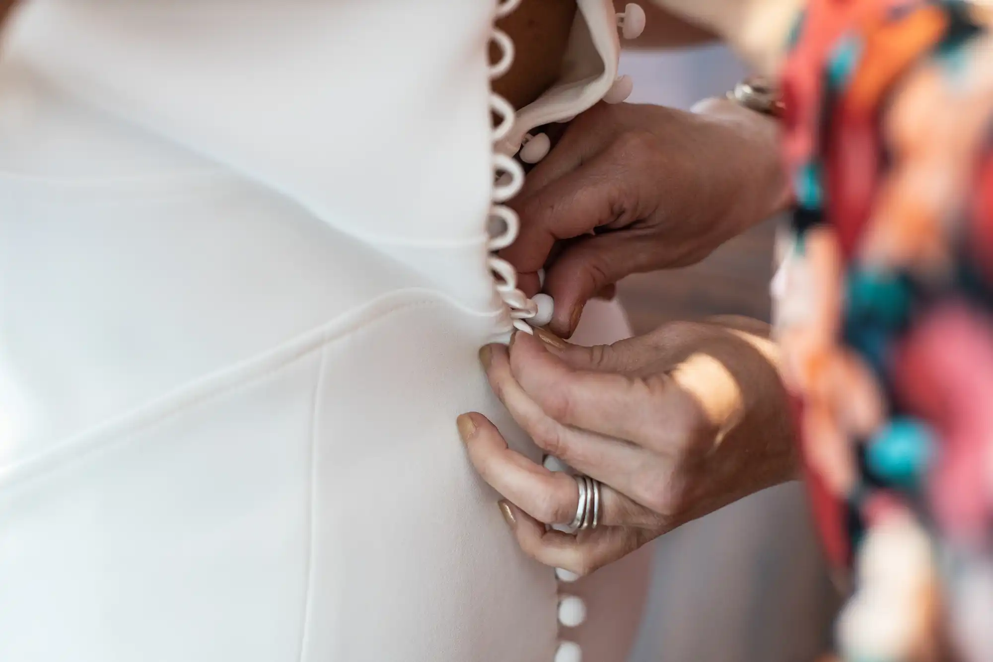 A close-up of a person's hands fastening the loop buttons on the back of a white wedding dress.
