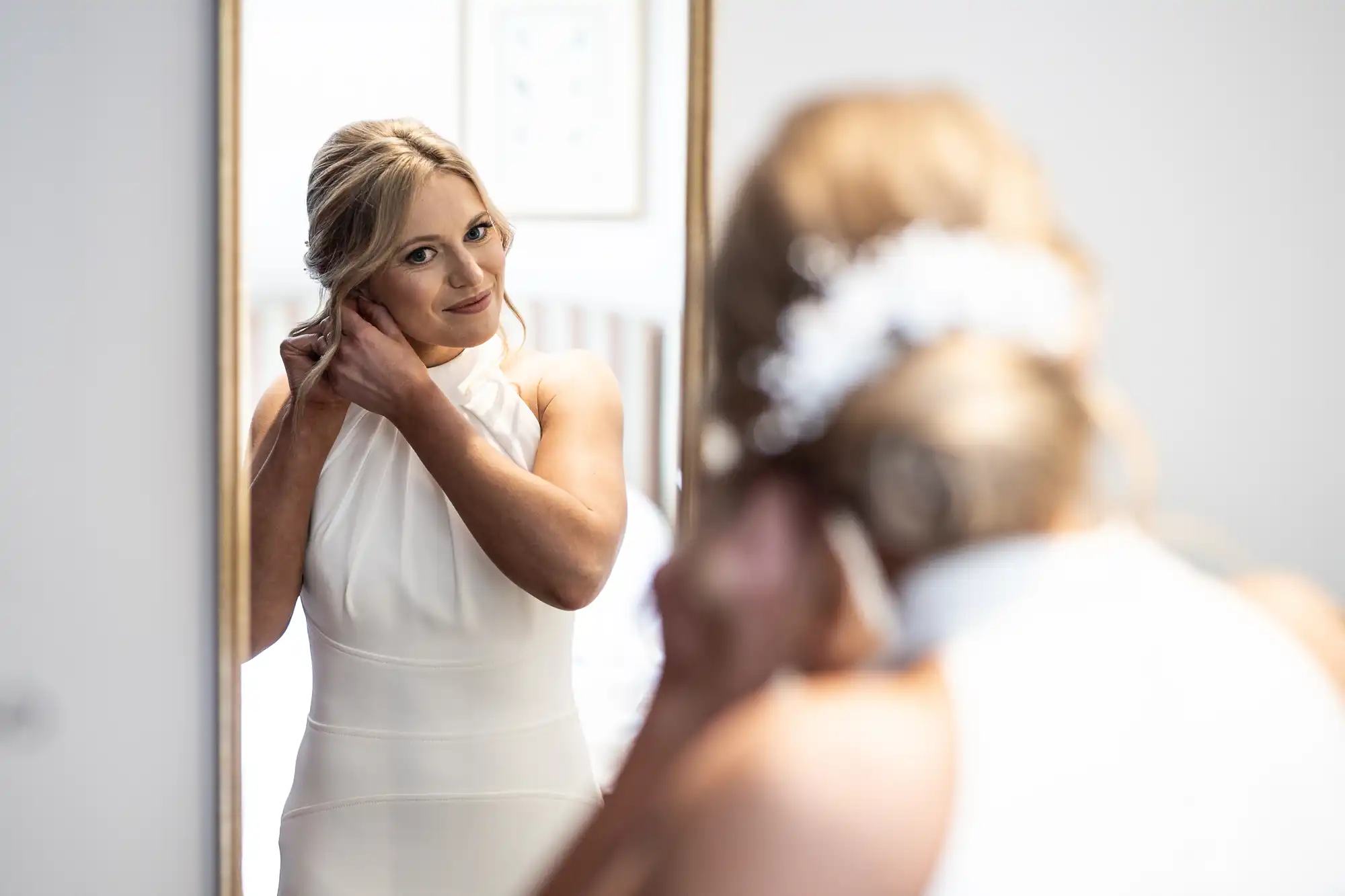 A bride in a white dress adjusts her earring while looking at her reflection in a mirror, smiling gently.