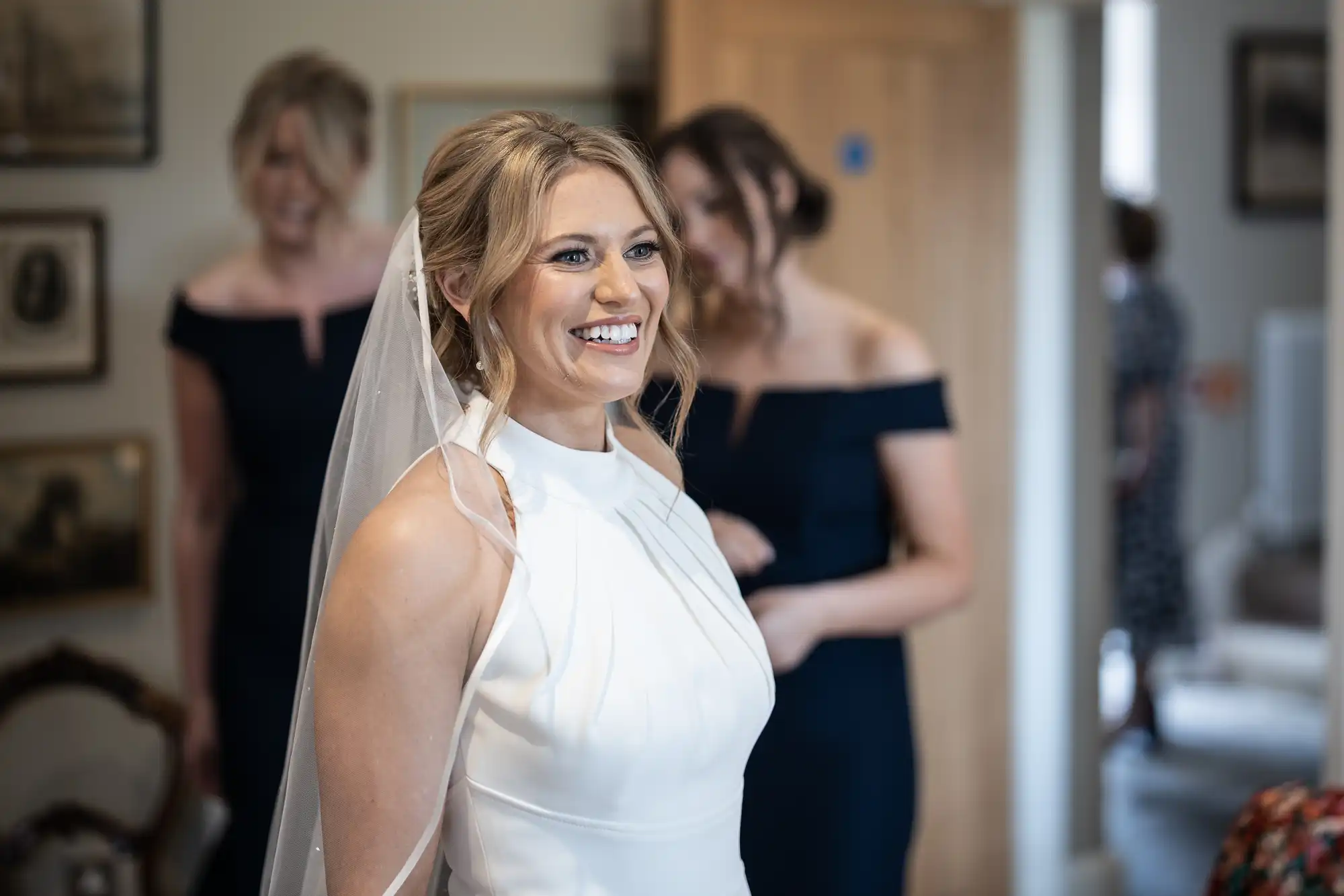 A smiling bride in a white halter-neck wedding dress and veil, with two women in navy dresses in the background.