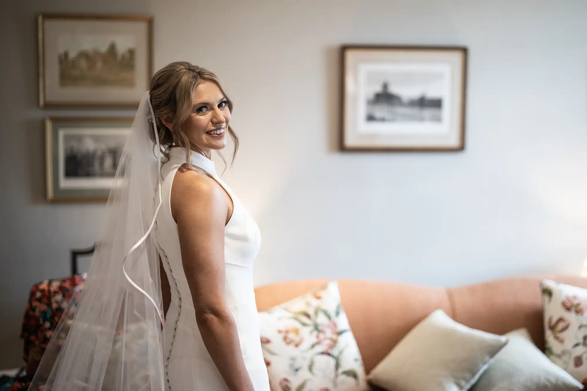 A bride in a white dress and veil smiles over her shoulder in a warmly lit room with elegant decor and paintings.