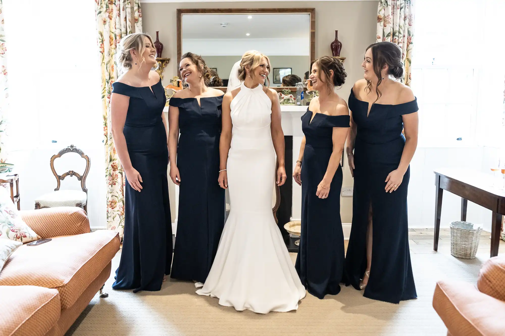 A bride in a white dress stands smiling with four bridesmaids in navy off-shoulder dresses in a warmly lit room.