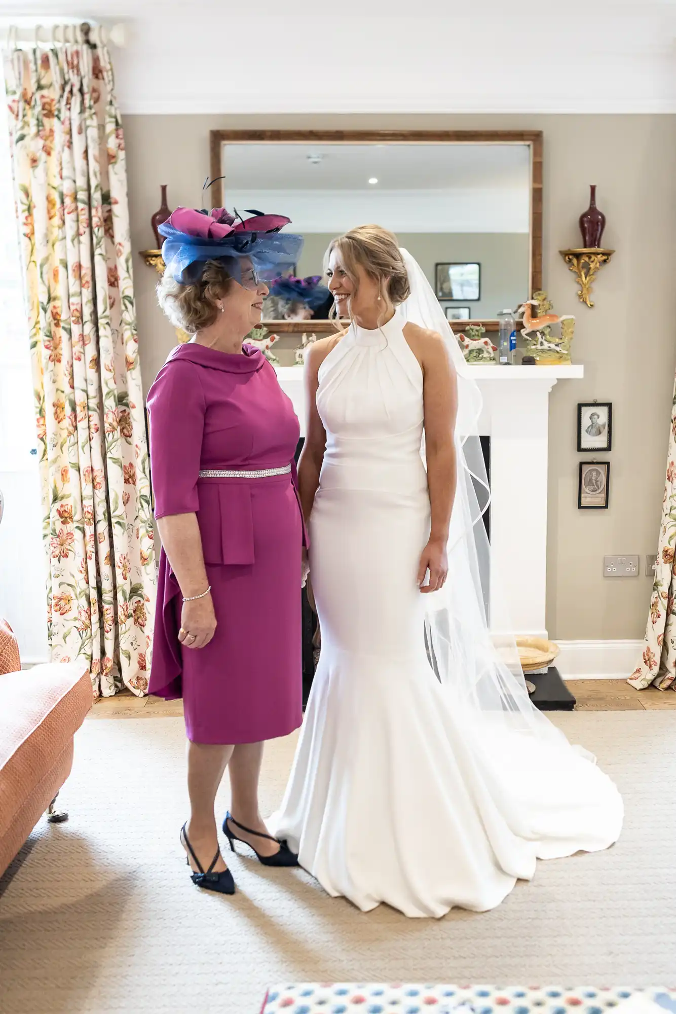 A bride in a white gown smiling at a woman in a magenta dress and blue hat in a well-lit room, both standing near a fireplace.