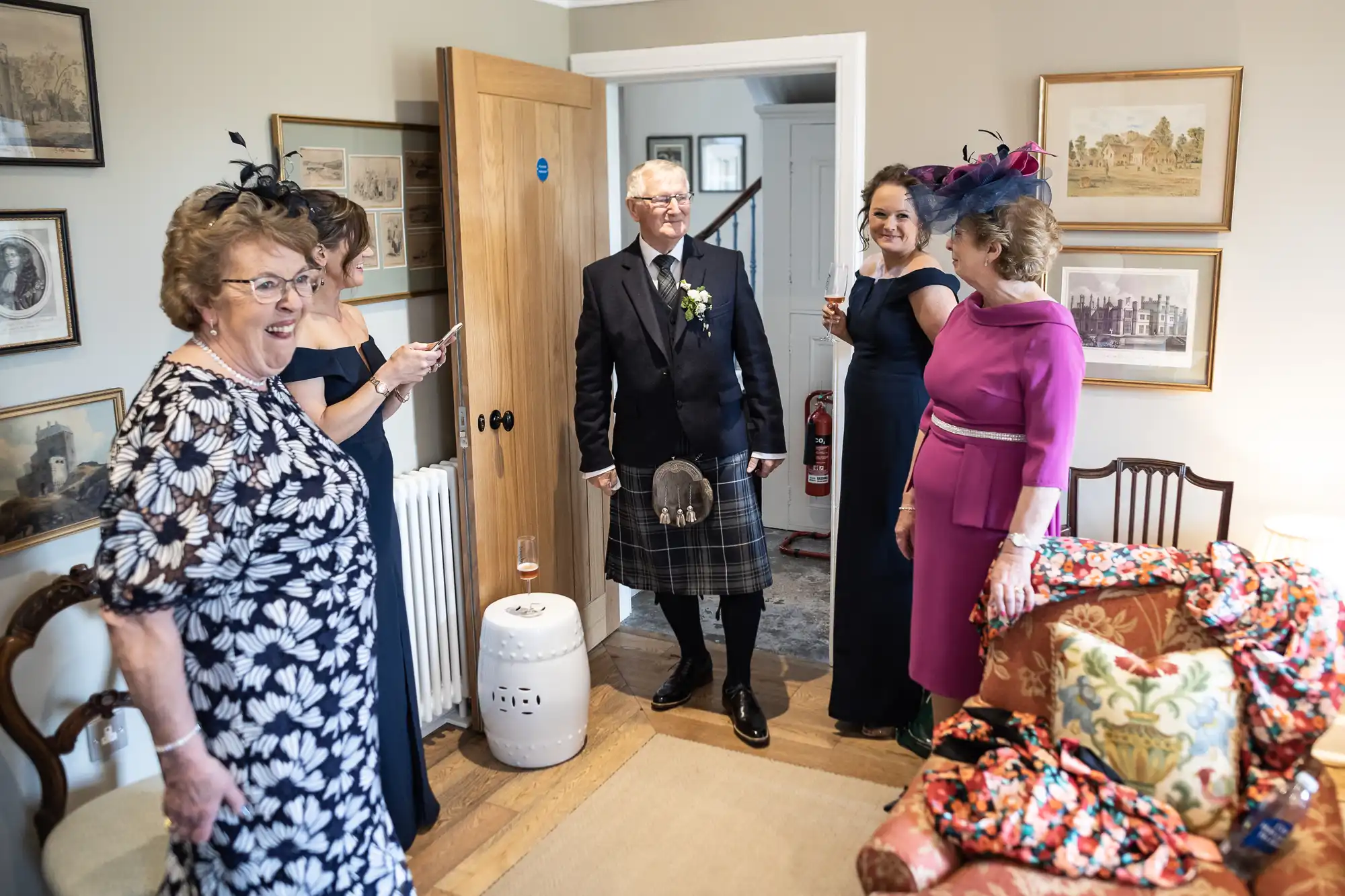 An elderly man in a kilt and three women in dresses and hats smiling and greeting each other in a cozy living room.