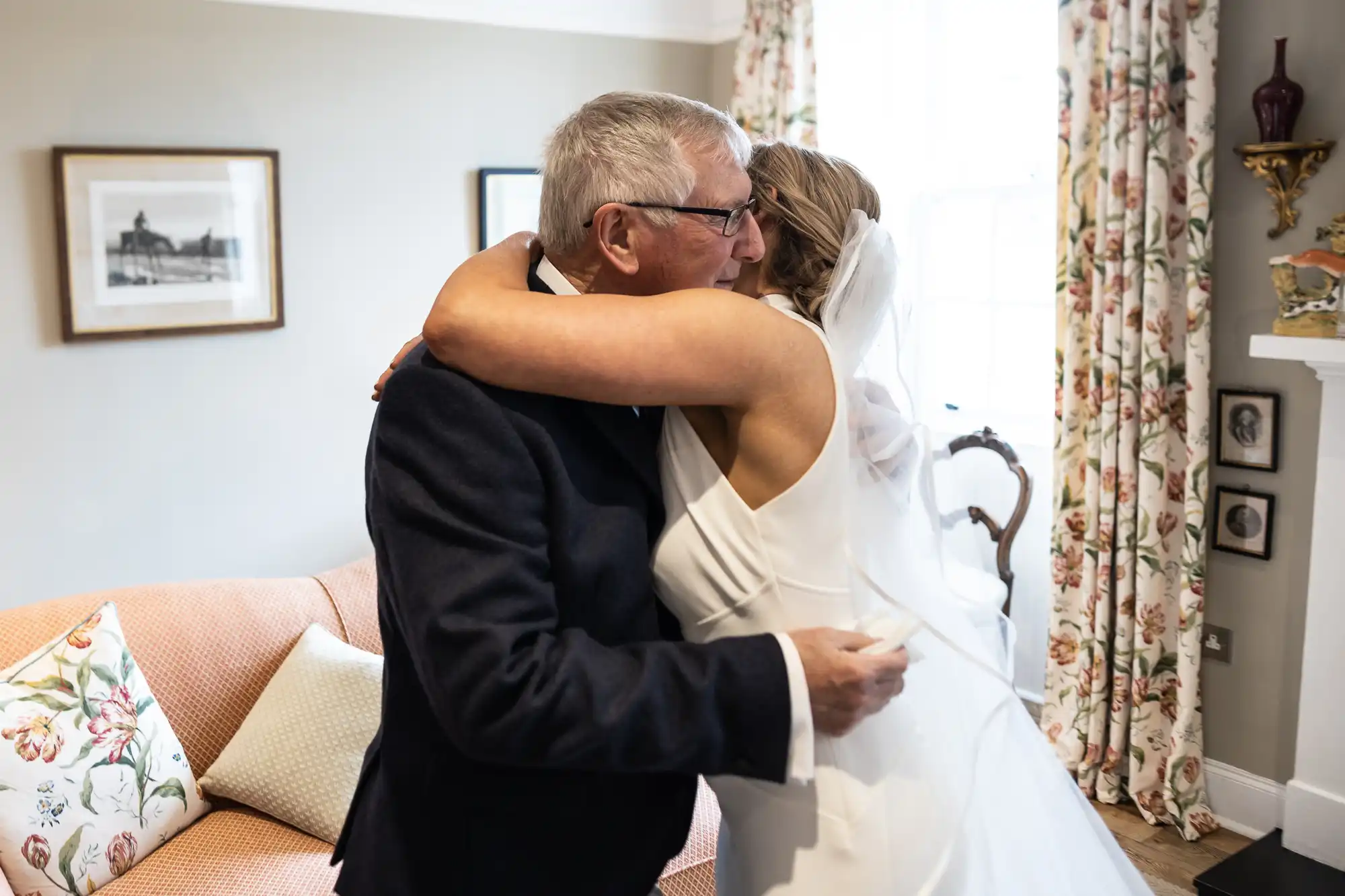 A bride in a white dress embracing an older man in a suit inside a room with elegant decor.
