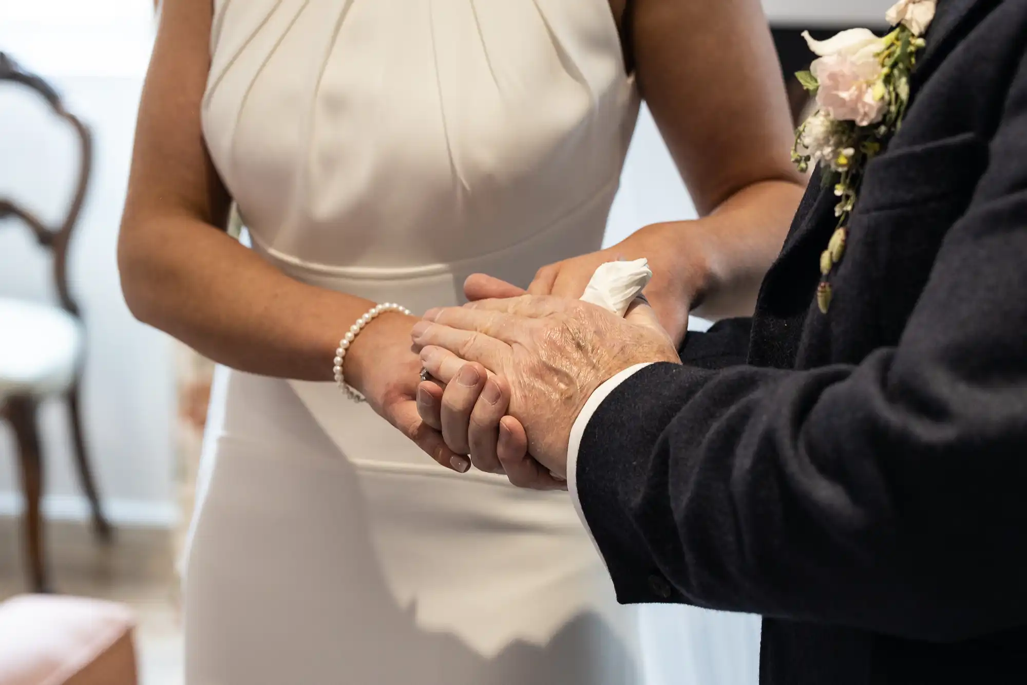 A close-up of a bride and groom holding hands during a wedding ceremony, showcasing their wedding attire and a corsage.