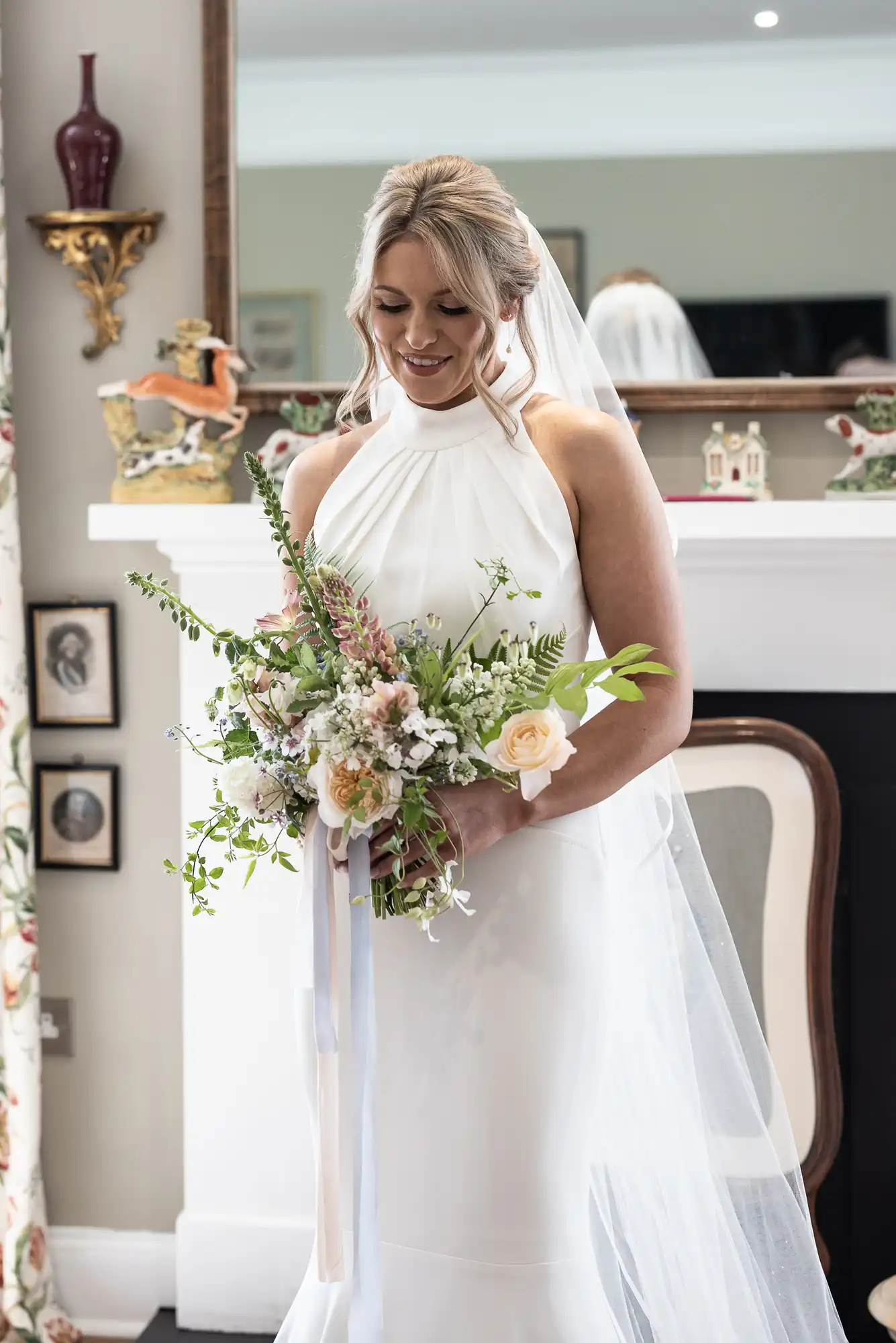 A bride in a white halter-neck gown holding a bouquet, smiling gently, indoors with decorative items in the background.