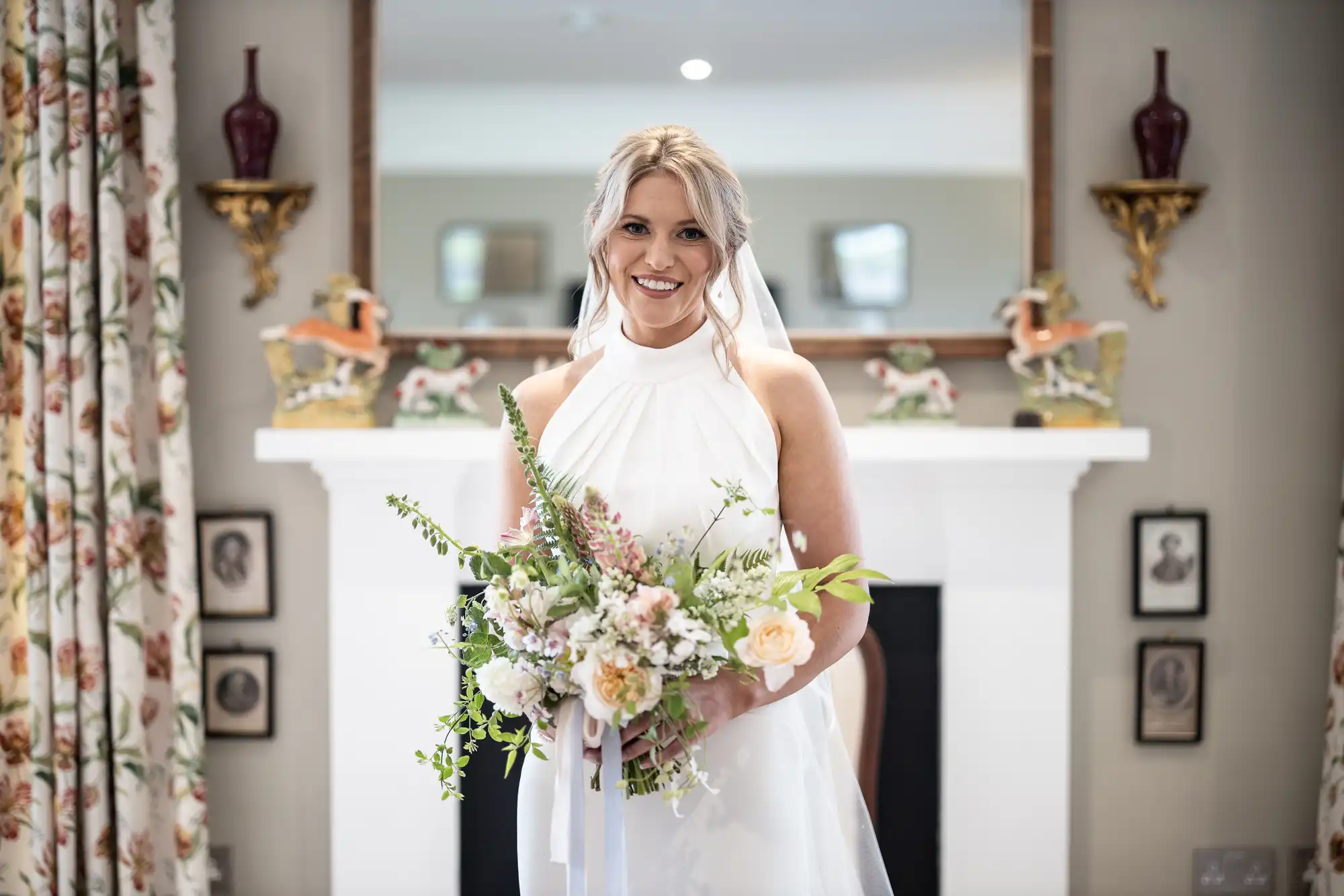 A bride smiling in a white dress and veil, holding a bouquet, standing in a room with elegant decor.