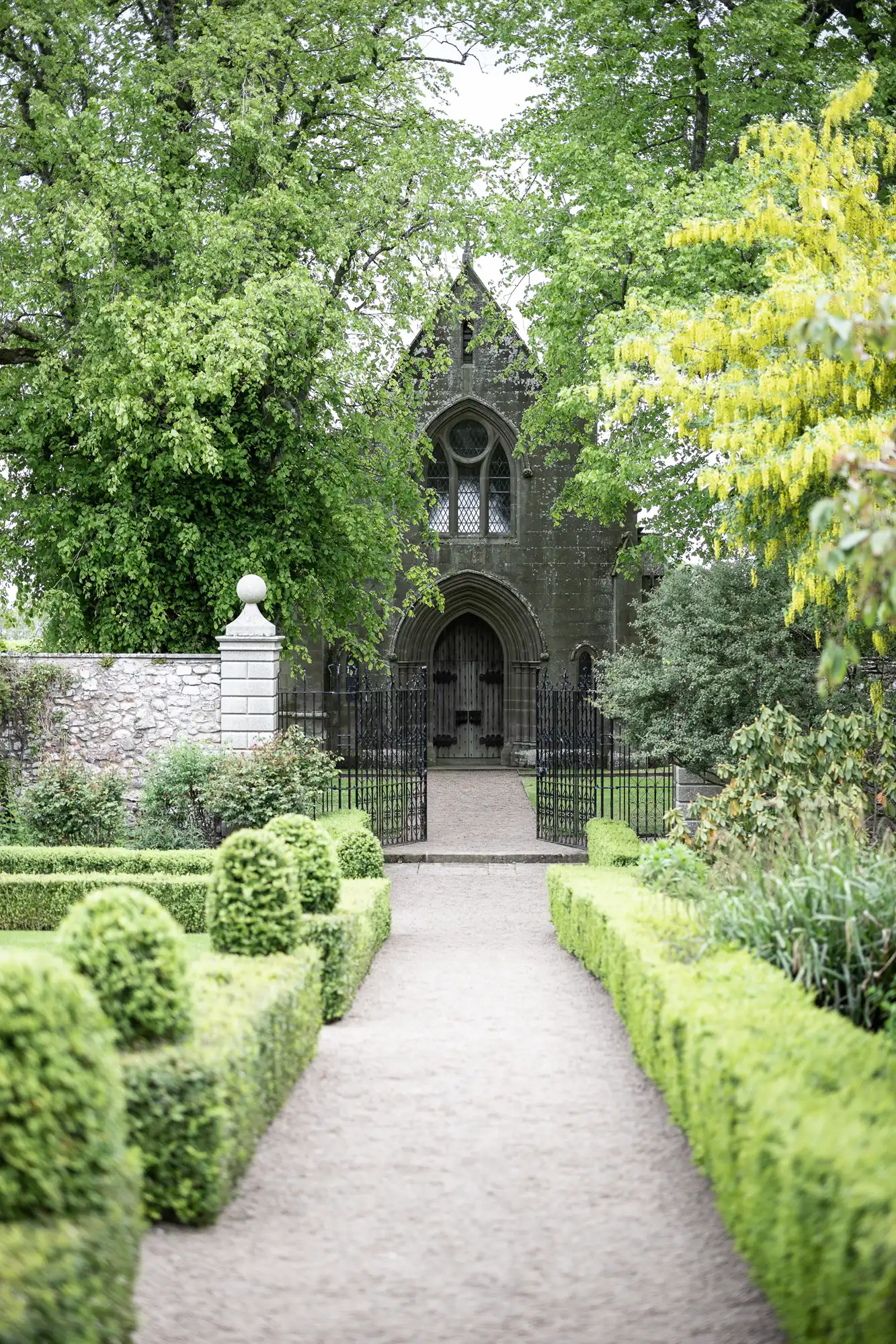 A landscaped garden path leads to a Gothic-style stone chapel surrounded by lush greenery and topiary hedges.