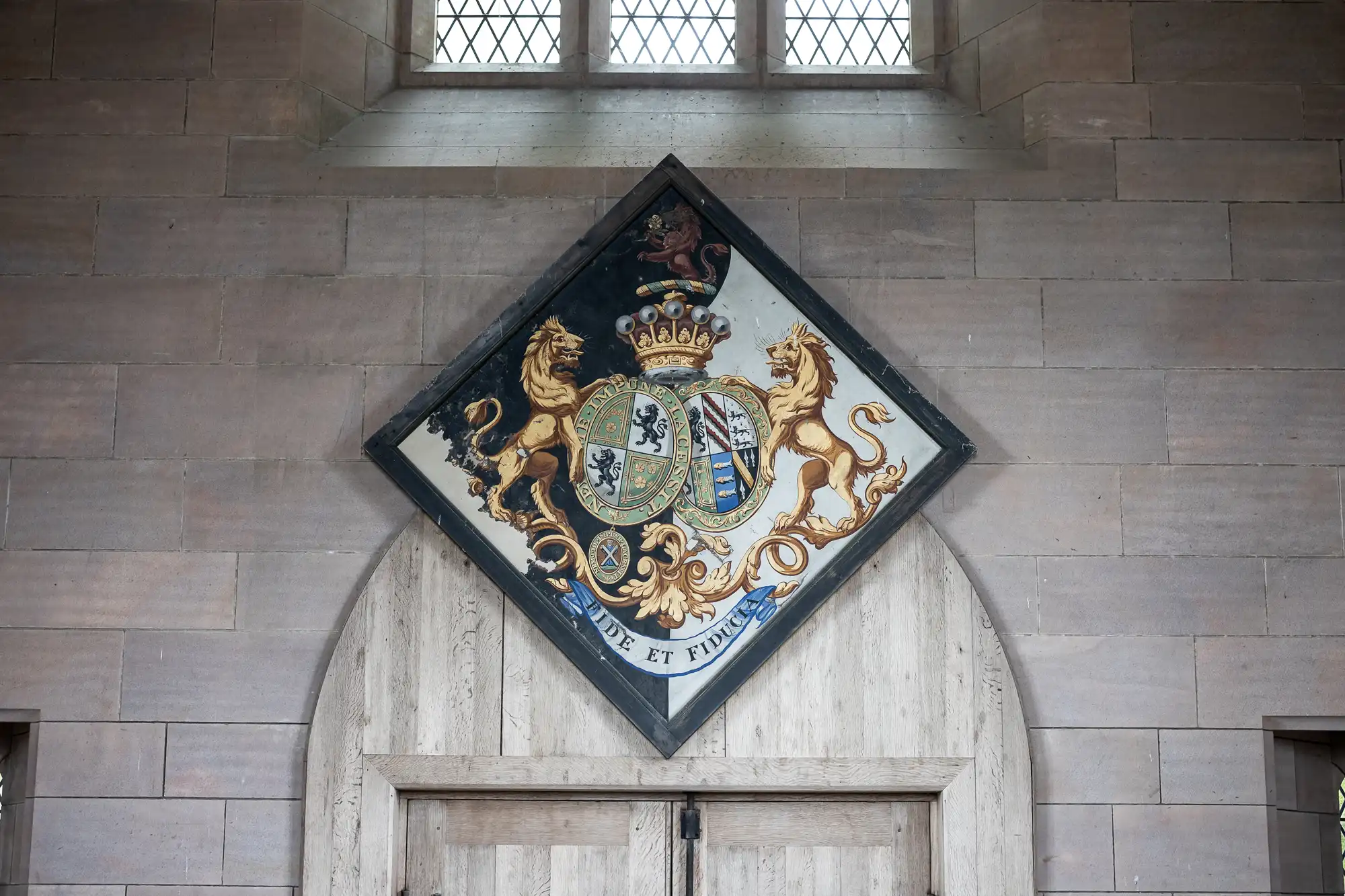 A heraldic coat of arms mounted above a wooden door inside a stone building, featuring a shield with a lion and a unicorn, and the motto "Dieu et mon droit.