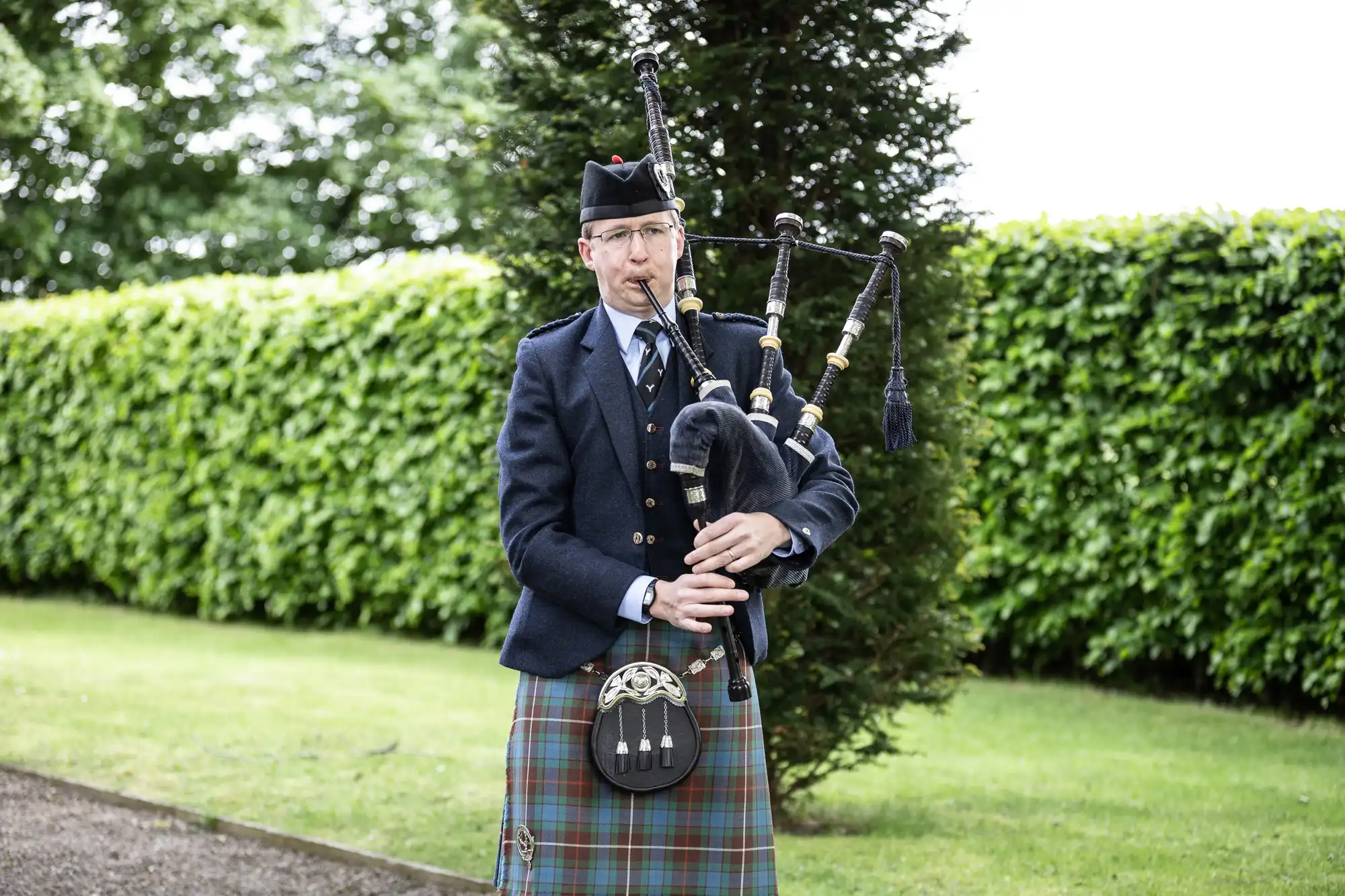 A man in traditional Scottish attire playing the bagpipes outdoors, surrounded by lush greenery.