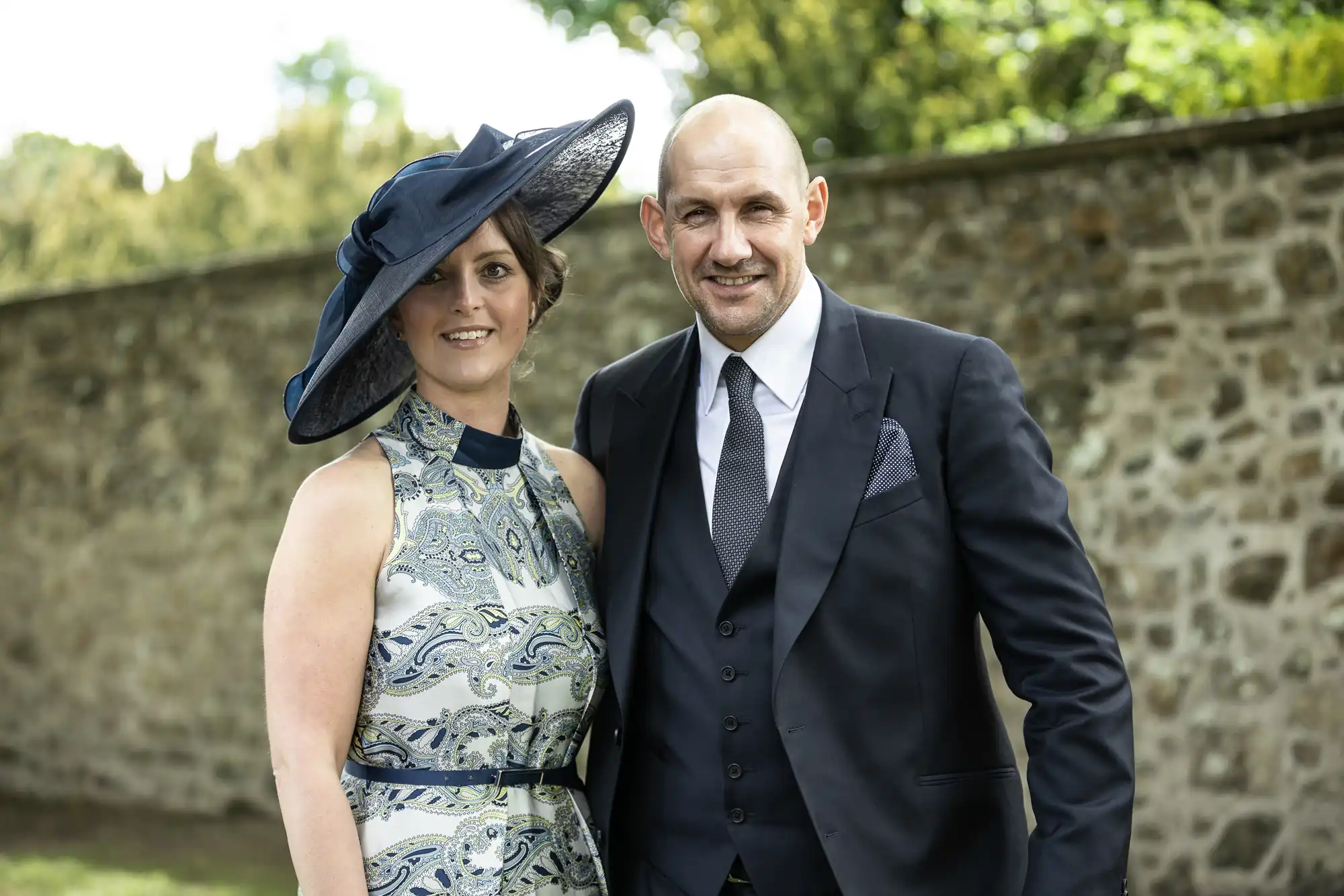 A well-dressed couple posing outdoors at a formal event, the woman in a patterned dress and wide-brimmed hat, and the bald man in a dark suit and tie.
