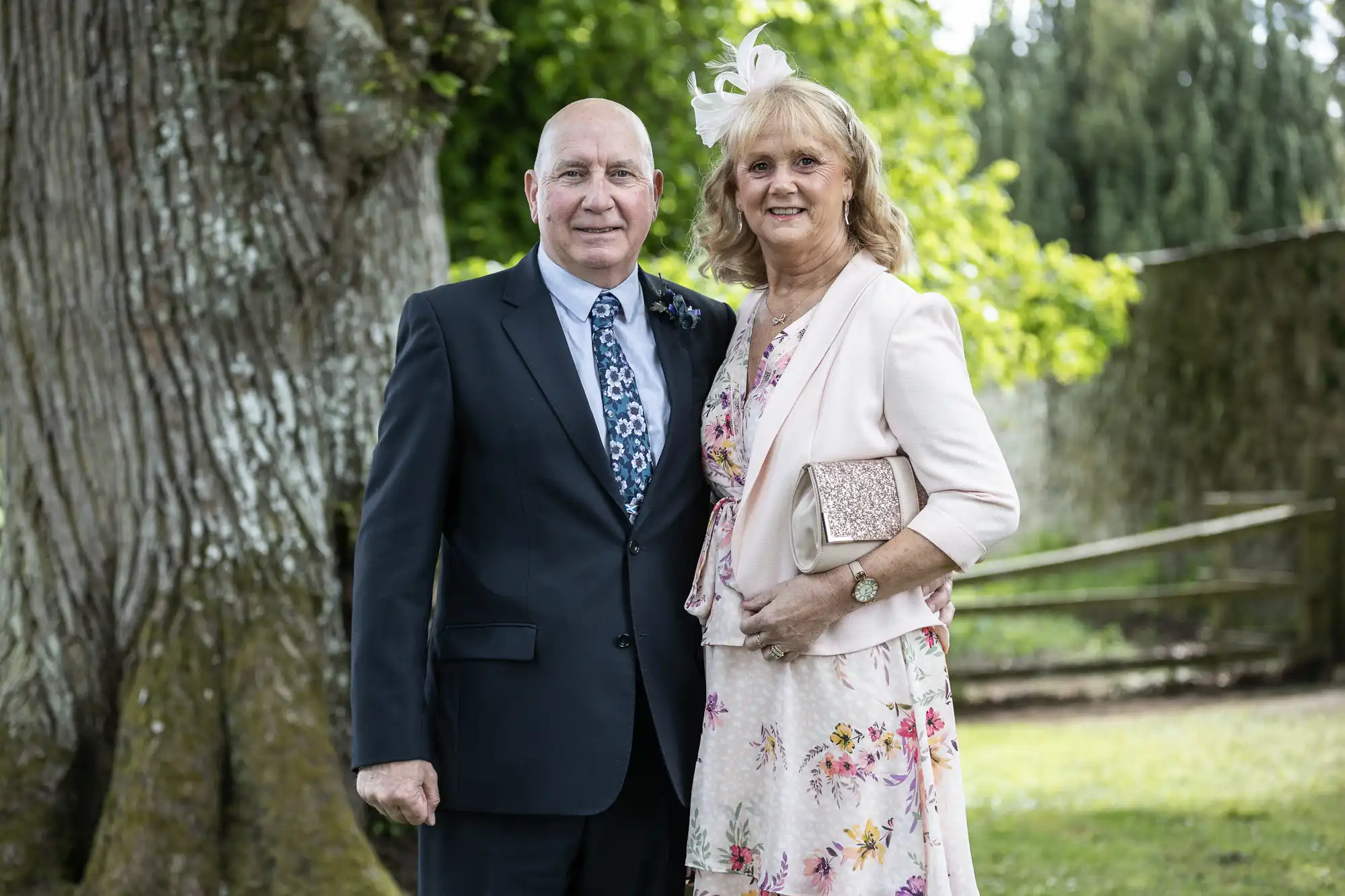 A mature couple dressed in formal wear, the man in a dark suit and the woman in a pink floral dress, posing together outdoors by a large tree.