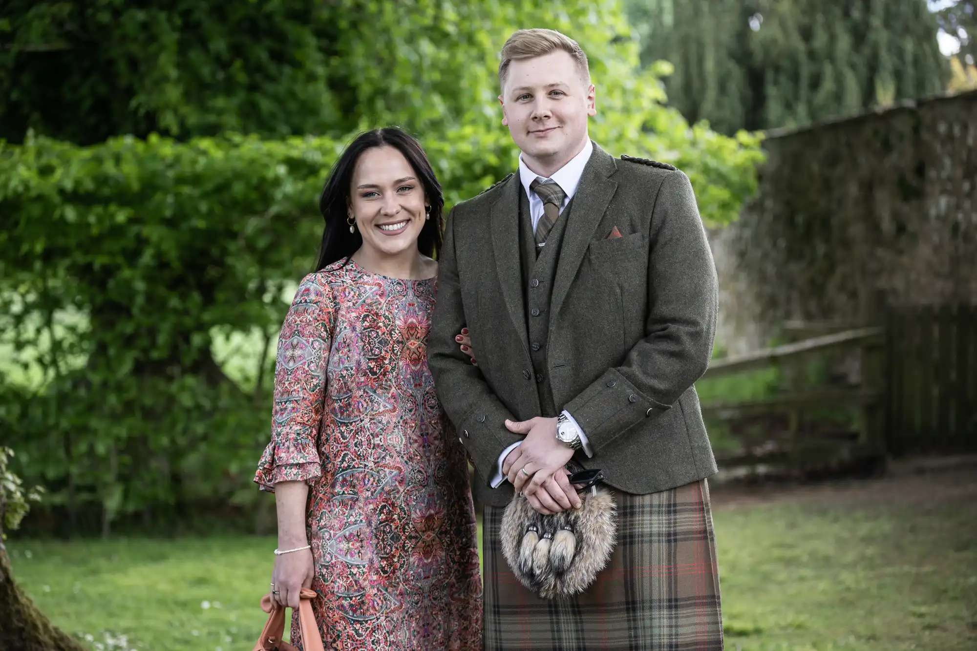 A woman in a floral dress and a man in a tartan kilt and tweed jacket stand side by side, smiling in a lush garden.