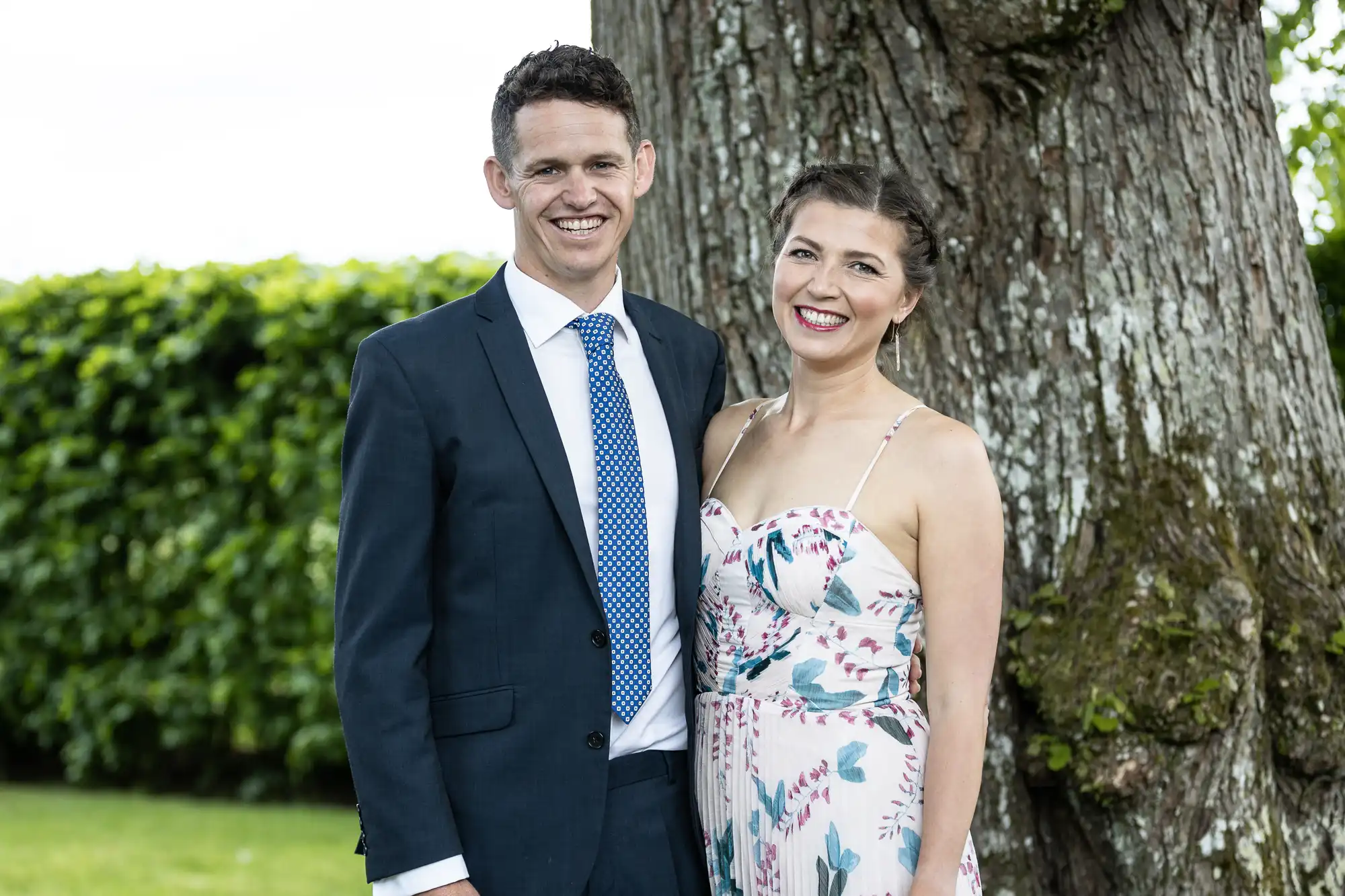 A smiling couple in formal attire, the man in a dark suit and the woman in a floral dress, standing by a large tree.