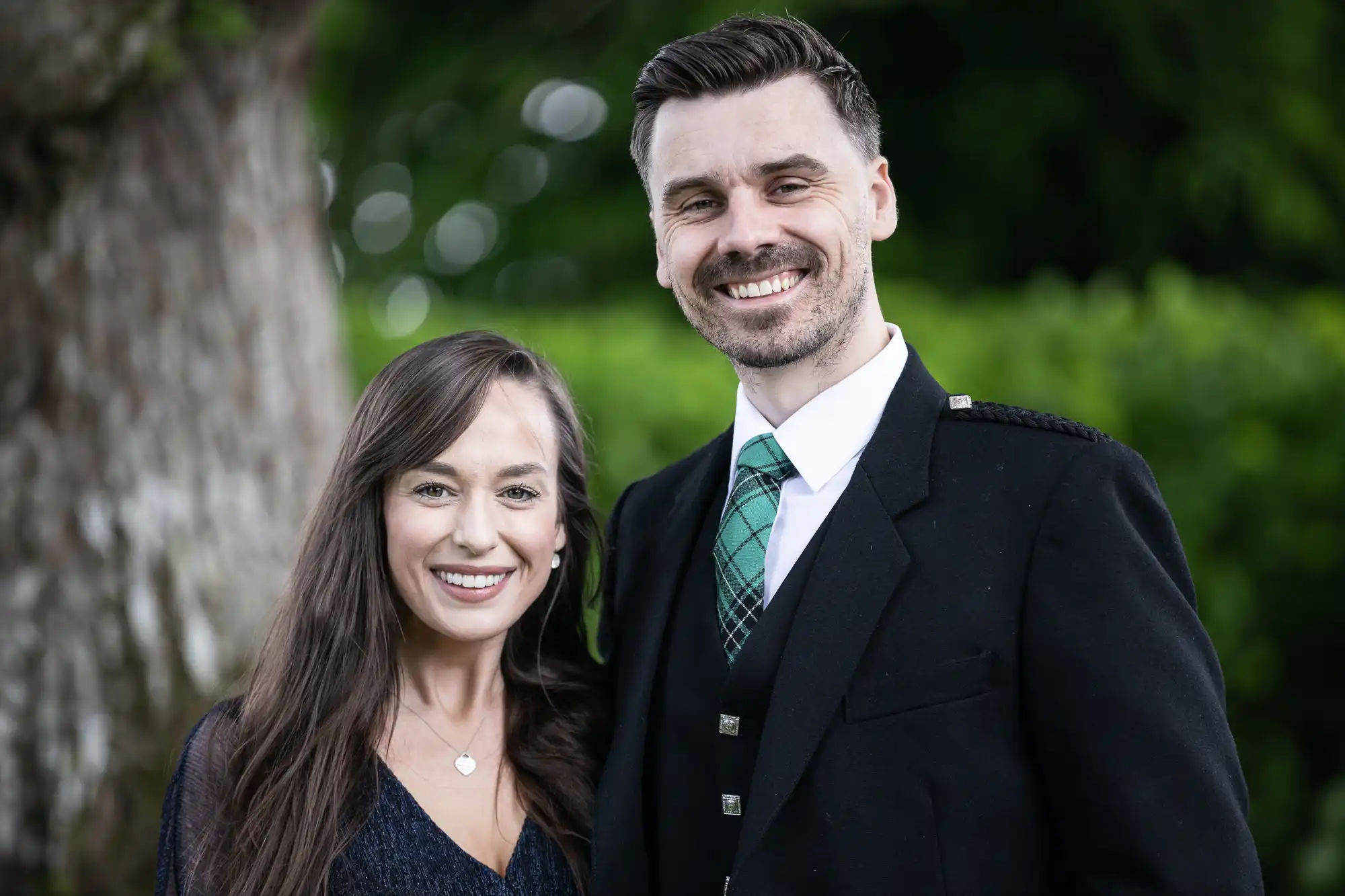A smiling couple posing outdoors, the man wearing a kilt and the woman in a dark dress, with a lush green background.