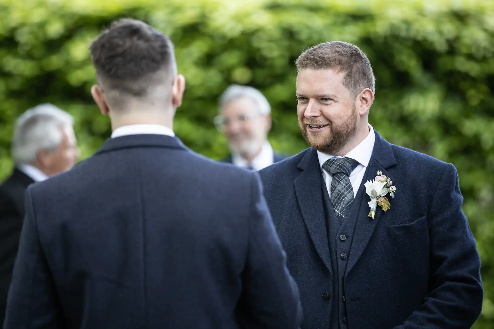 A groom in a suit with a boutonniere smiles at another man in a formal setting, with older guests in the blurred background.