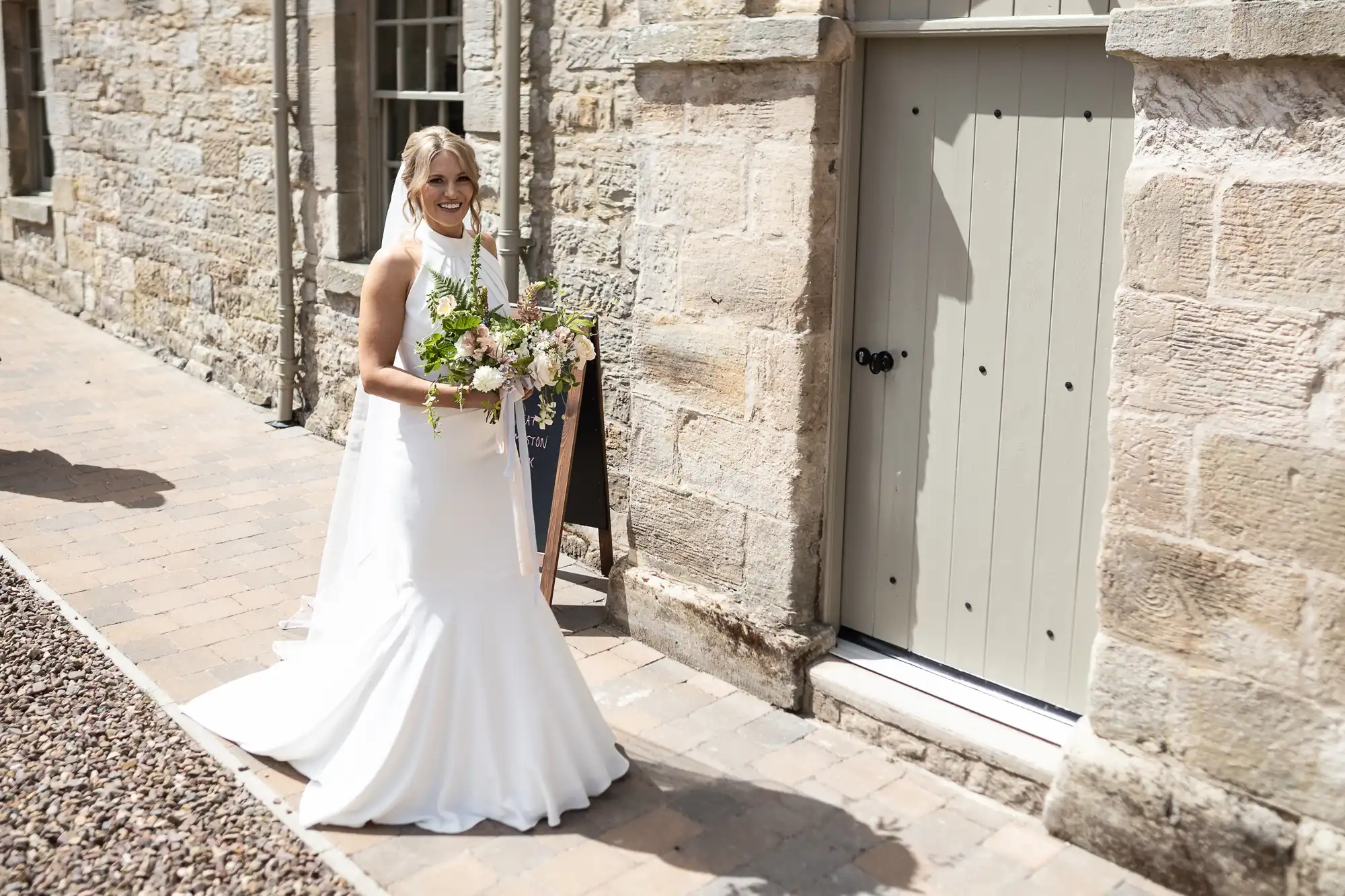A bride in a white dress holding a bouquet, smiling next to a stone building with a pale green door.