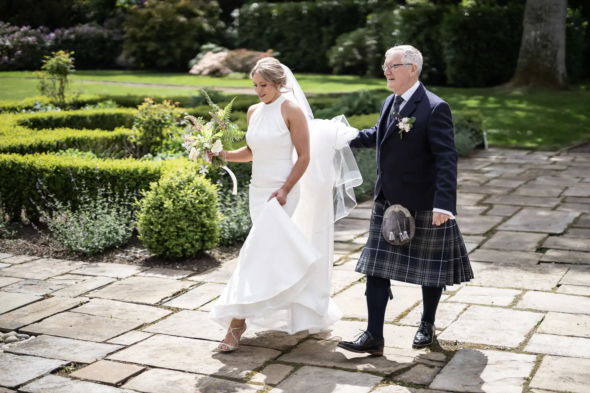 A bride and her father walking in a garden, the father wearing a kilt and the bride holding a bouquet.
