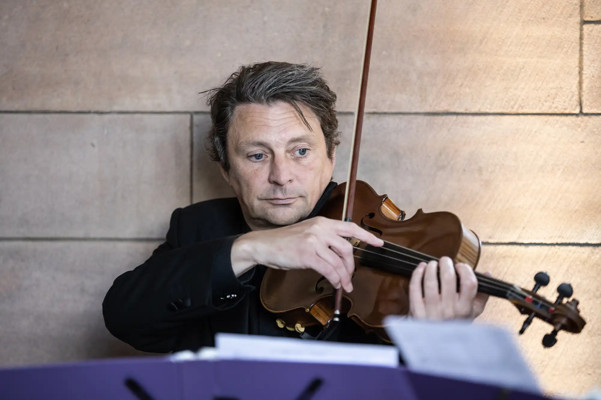 Man in a black suit playing a violin intently, seated next to sheet music.