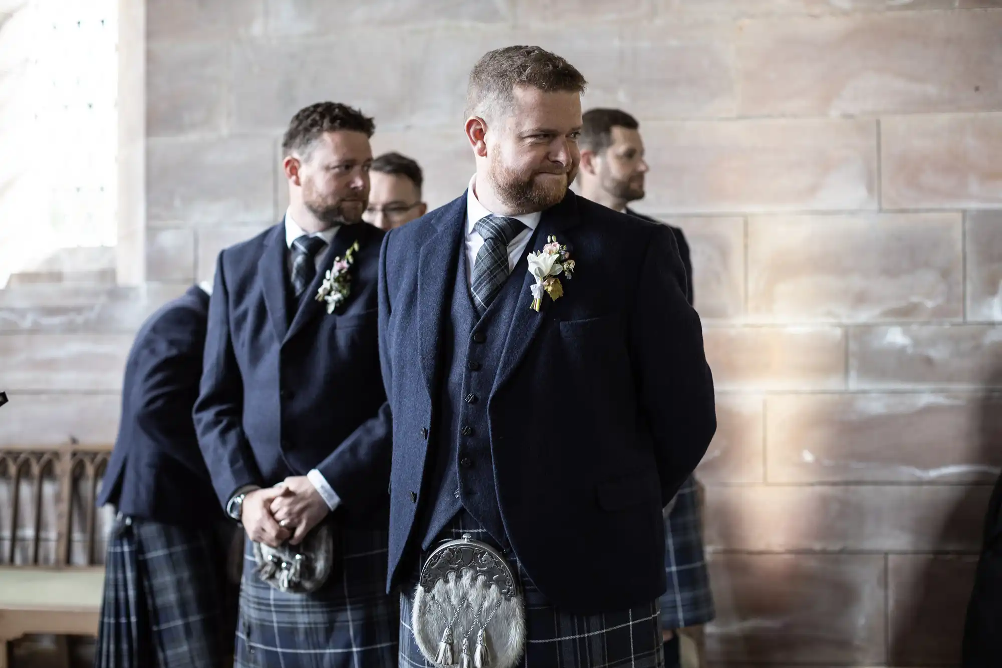 A group of men in traditional Scottish attire, including kilts and jackets, stand solemnly in a room with a stone wall background. One man in the foreground smiles slightly.