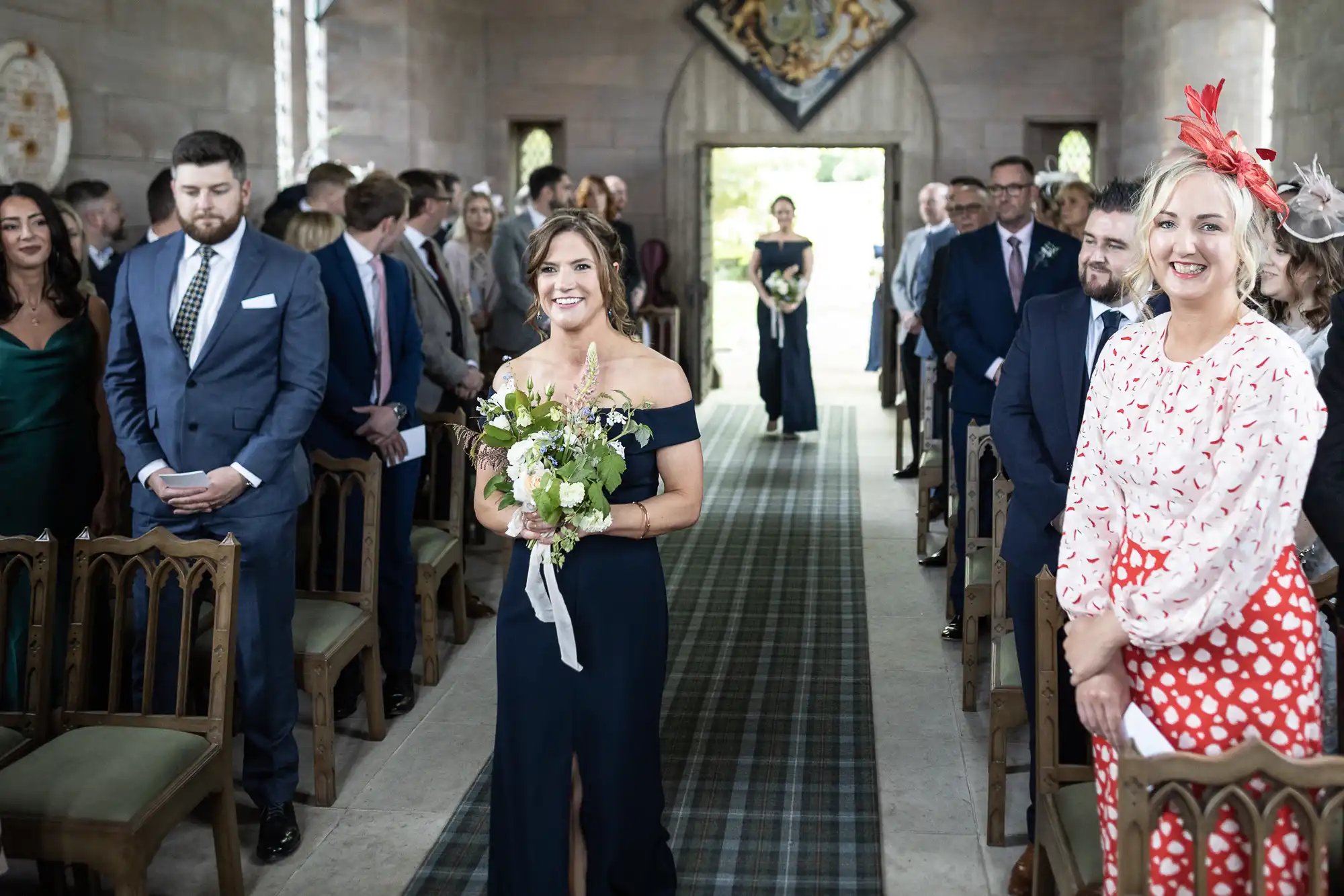 Bridesmaid smiling and walking down the aisle in a church, holding a bouquet, with guests standing and watching.