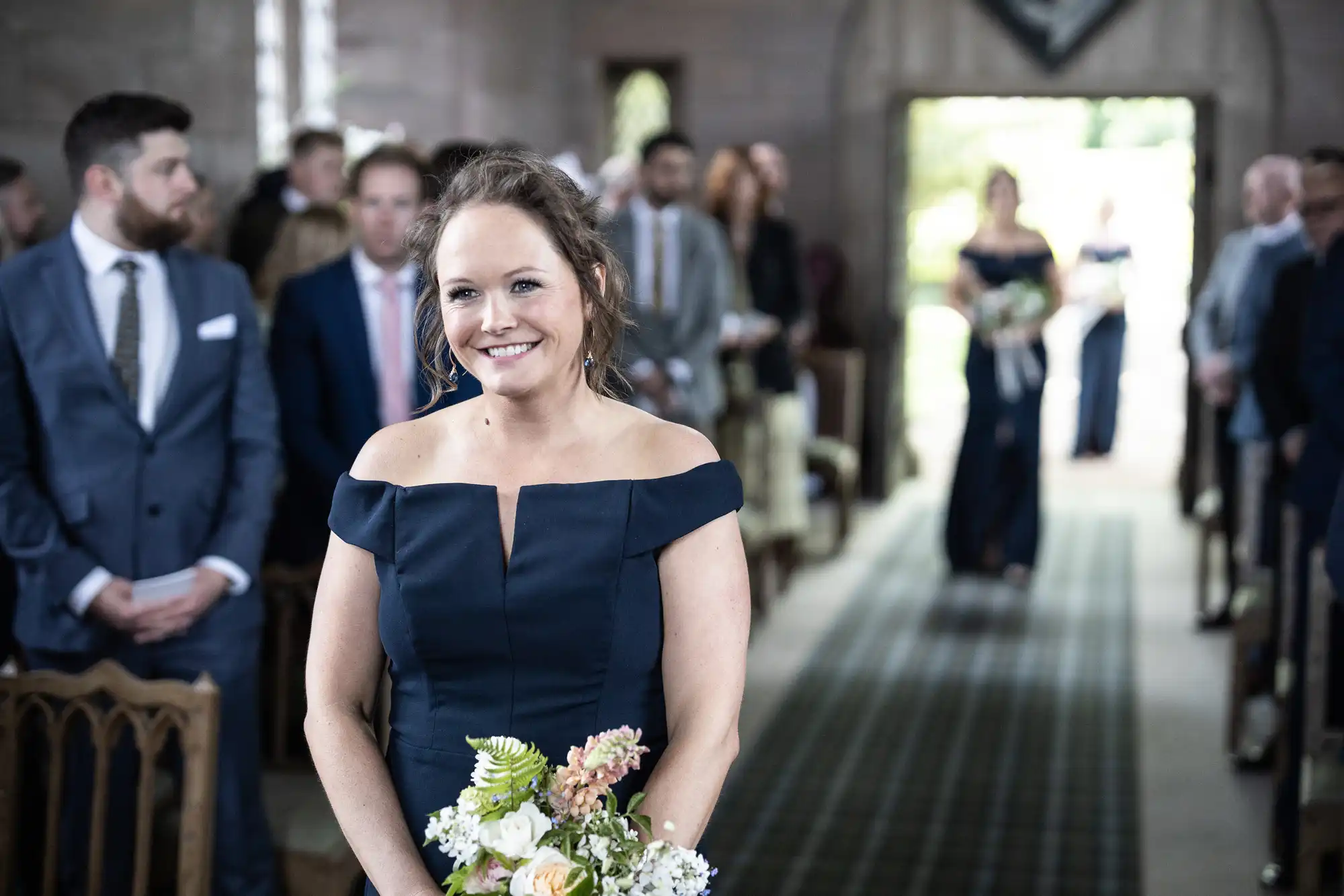 A woman in a navy off-shoulder dress smiling with a bouquet, standing in a church aisle with guests in the background.